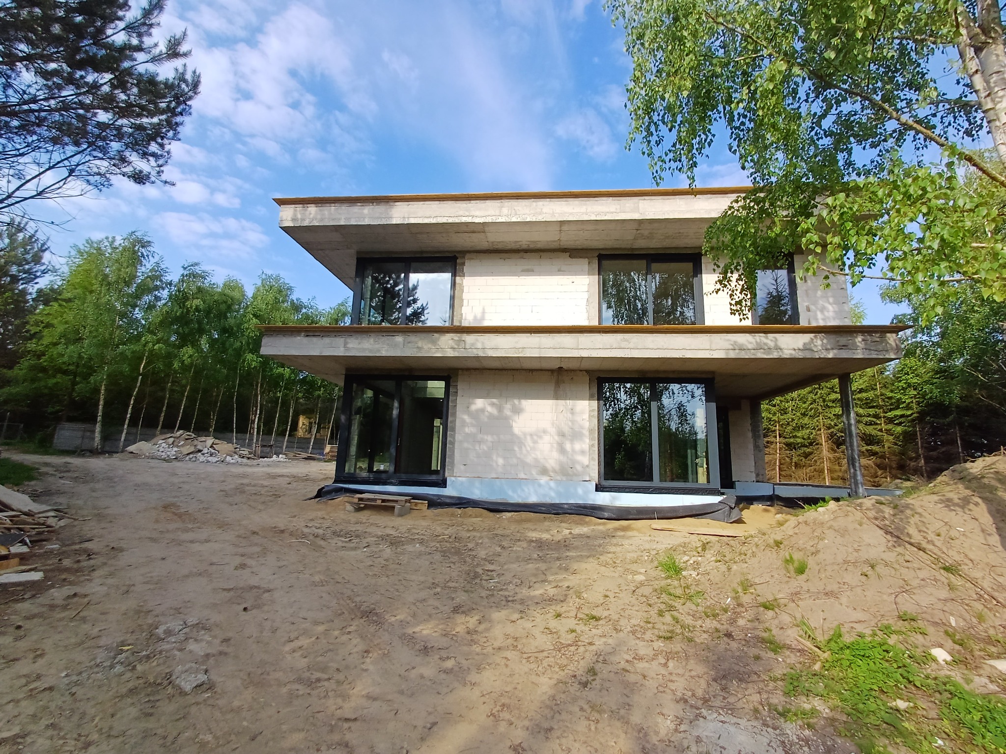 Surowy, dwukondygnacyjny budynek w trakcie budowy, z widocznymi blokami i betonowymi elementami konstrukcyjnymi, duże okna z czarnymi ramami, otoczony drzewami i niezagospodarowanym terenem.