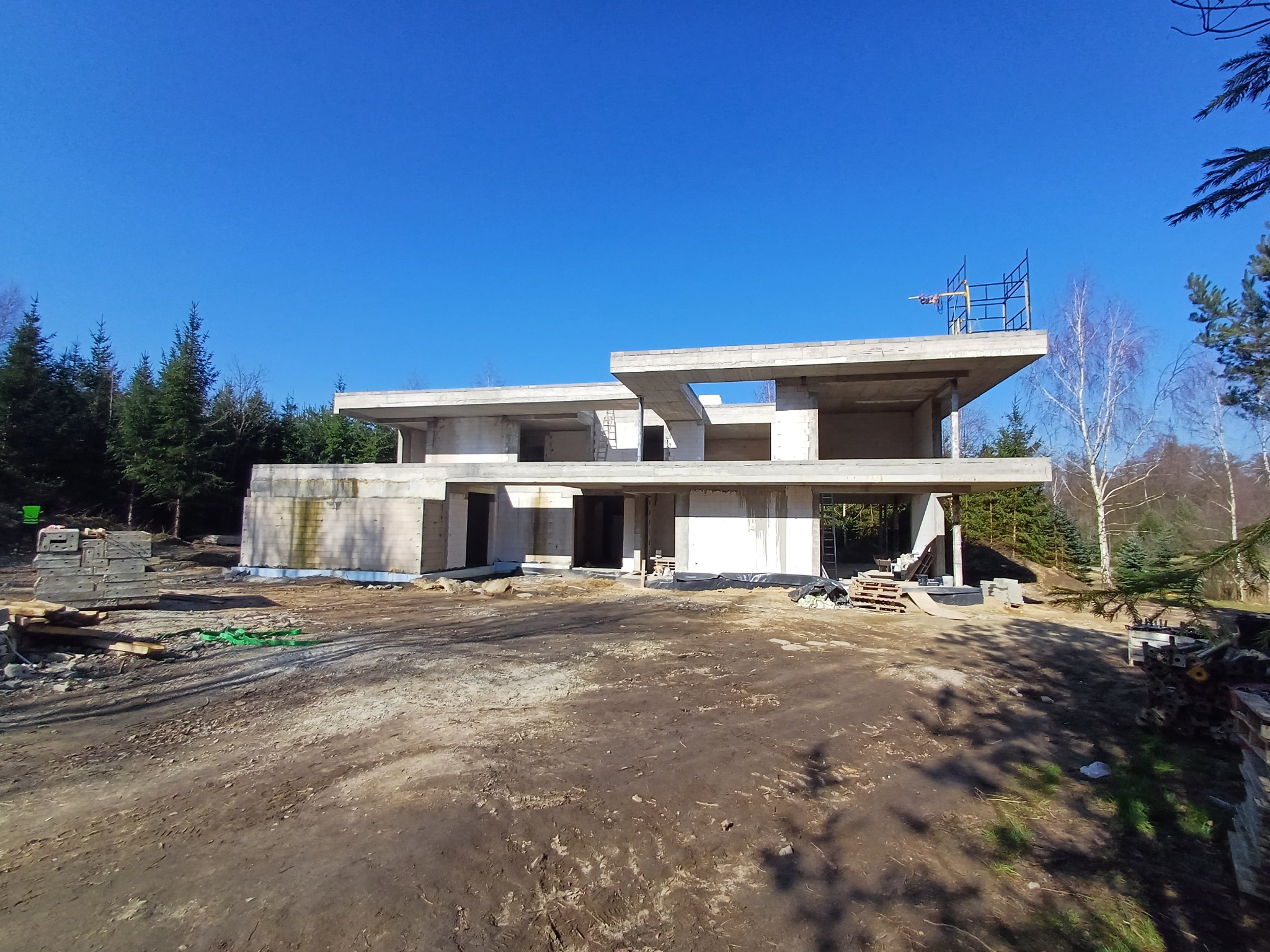 Surowy, dwukondygnacyjny budynek żelbetowy w trakcie budowy, z widocznymi rusztowaniami na dachu i stertą materiałów budowlanych na placu, otoczony drzewami i błękitnym niebem.