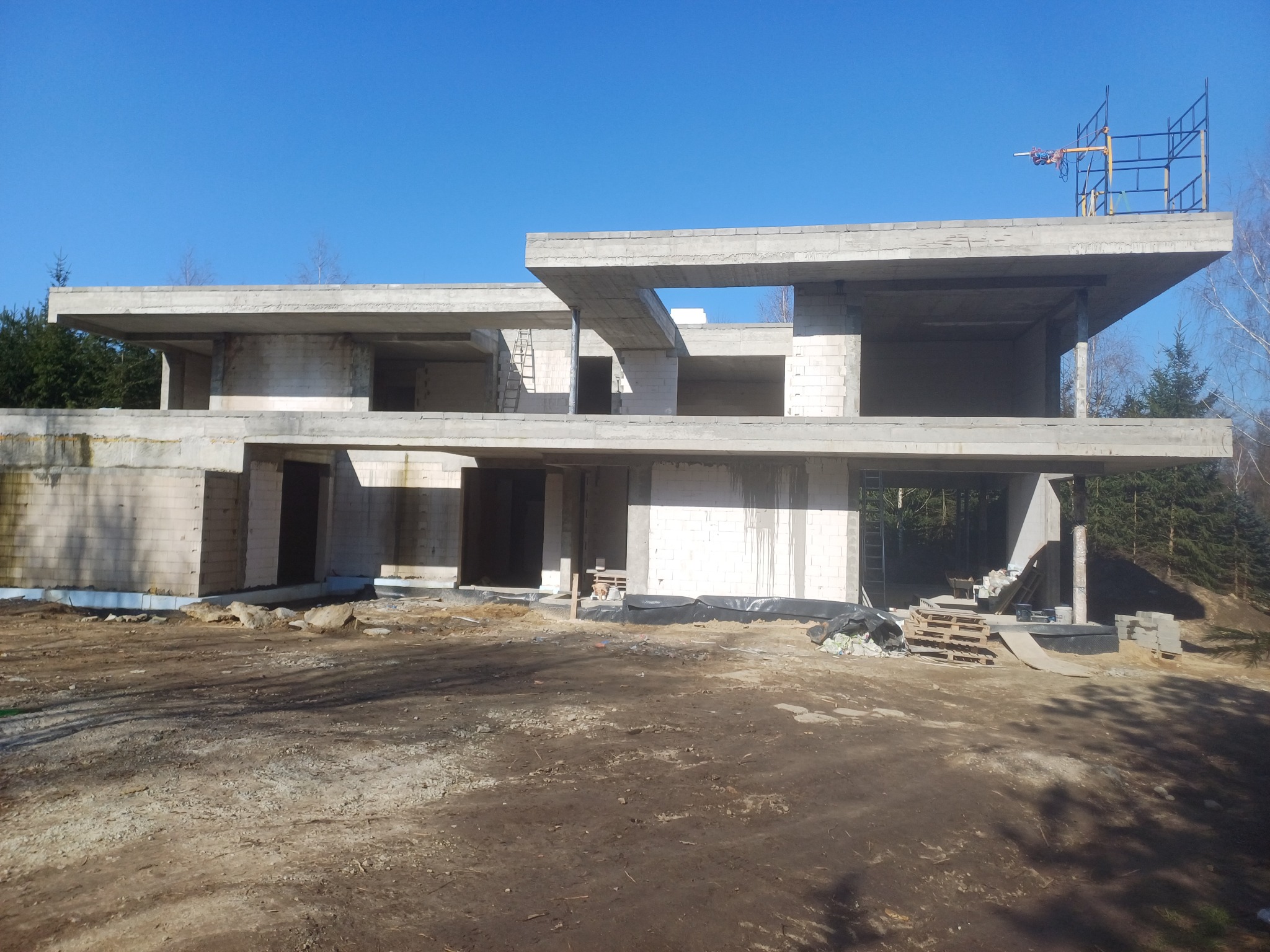 Surowy stan budynku żelbetowego w trakcie budowy, widoczne ściany z bloczków, szalunki stropowe, rusztowanie na dachu, teren budowy z ziemią i paletami.