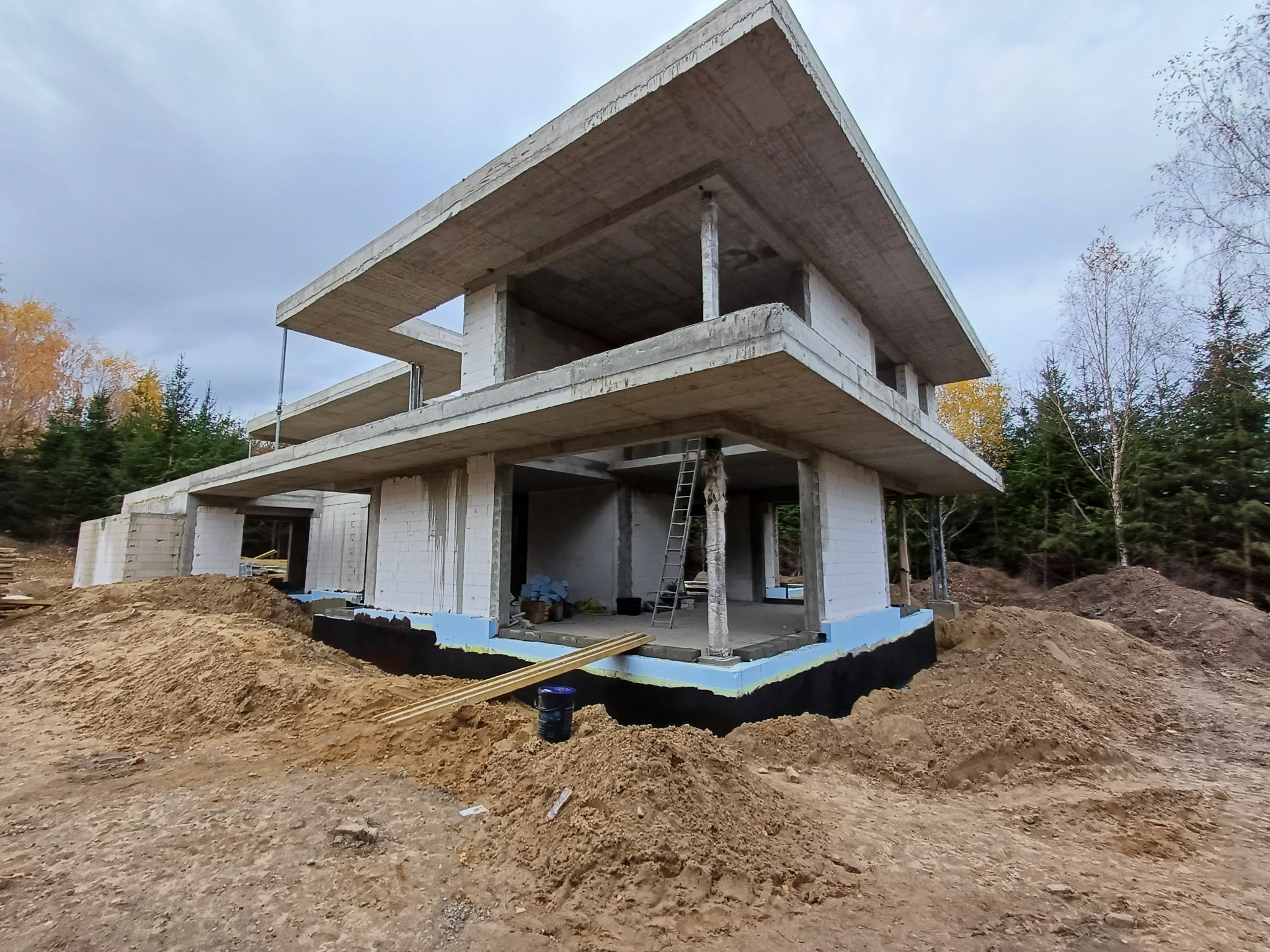Surowy stan dwukondygnacyjnego domu z betonu, z widocznymi ścianami z bloczków, izolacją fundamentów w kolorze niebieskim i czarnym, oraz stertą ziemi wokół, drabina oparta o filar.