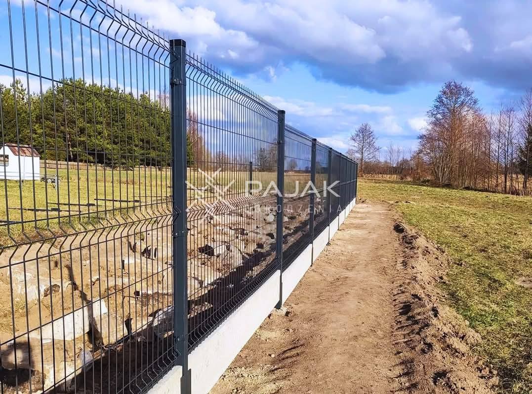 Długi, grafitowy system ogrodzeniowy z paneli na betonowej podmurówce, widoczny w krajobrazie wiejskim z polami i budynkiem w tle, z logo firmy na przęśle.