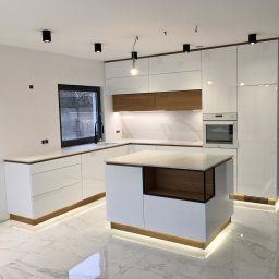 Pracownia m2 - Nowoczesna, biała kuchnia z wyspą i marmurową podłogą, oświetlenie LED pod szafkami, drewniane akcenty i czarne detale.