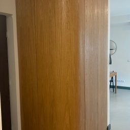 Pracownia m2 - Drewniana zabudowa meblowa z zaokrąglonym narożnikiem, dopasowana do wnęki w przedpokoju, widoczny fragment drzwi i wejście do innego pomieszczenia w tle