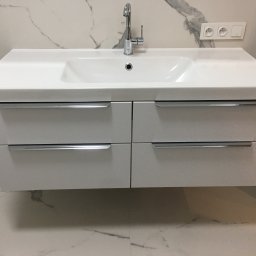 Pracownia m2 - Biała umywalka z czterema szufladami i chromowanymi uchwytami, zamontowana na ścianie wyłożonej marmurowymi płytkami z szarymi żyłami, bateria umywalkowa w kolorze chromu.