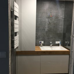 Pracownia m2 - Nowoczesna łazienka w trakcie wykańczania: szare płytki, białe meble łazienkowe z drewnianym blatem, grzejnik drabinkowy i widoczne przewody elektryczne.