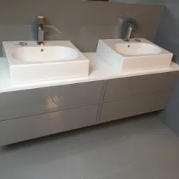 Dwie białe, prostokątne umywalki nablatowe z chromowanymi bateriami, osadzone na szarej szafce z czterema szufladami w nowoczesnej łazience z szarymi płytkami.