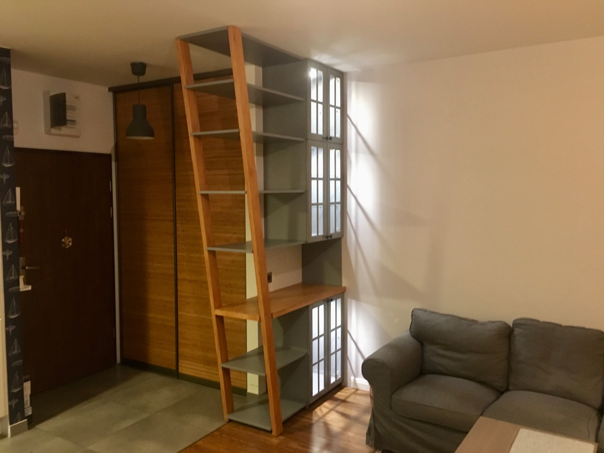 Regał na książki z drabinką, szary z drewnianymi elementami, w tle brązowa szafa przesuwna i szara sofa w salonie.