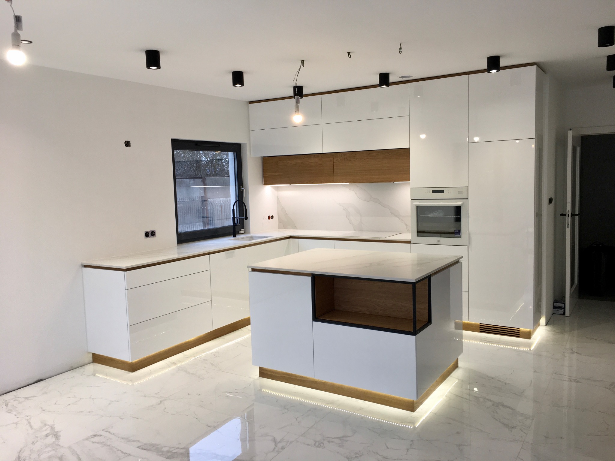Nowoczesna, biała kuchnia z wyspą i marmurową podłogą, oświetlenie LED pod szafkami, drewniane akcenty i czarne detale.