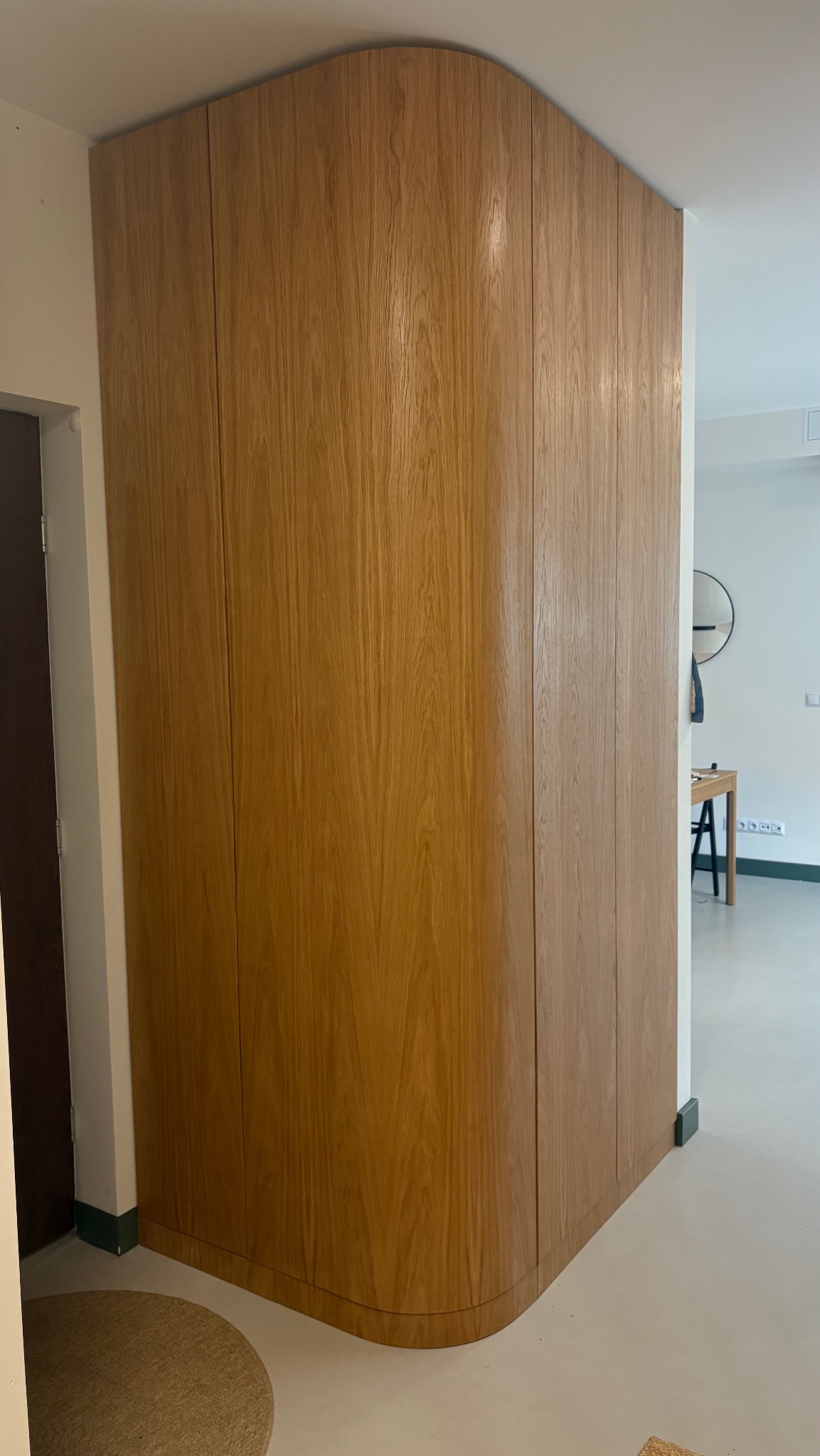 Drewniana zabudowa meblowa z zaokrąglonym narożnikiem, dopasowana do wnęki w przedpokoju, widoczny fragment drzwi i wejście do innego pomieszczenia w tle