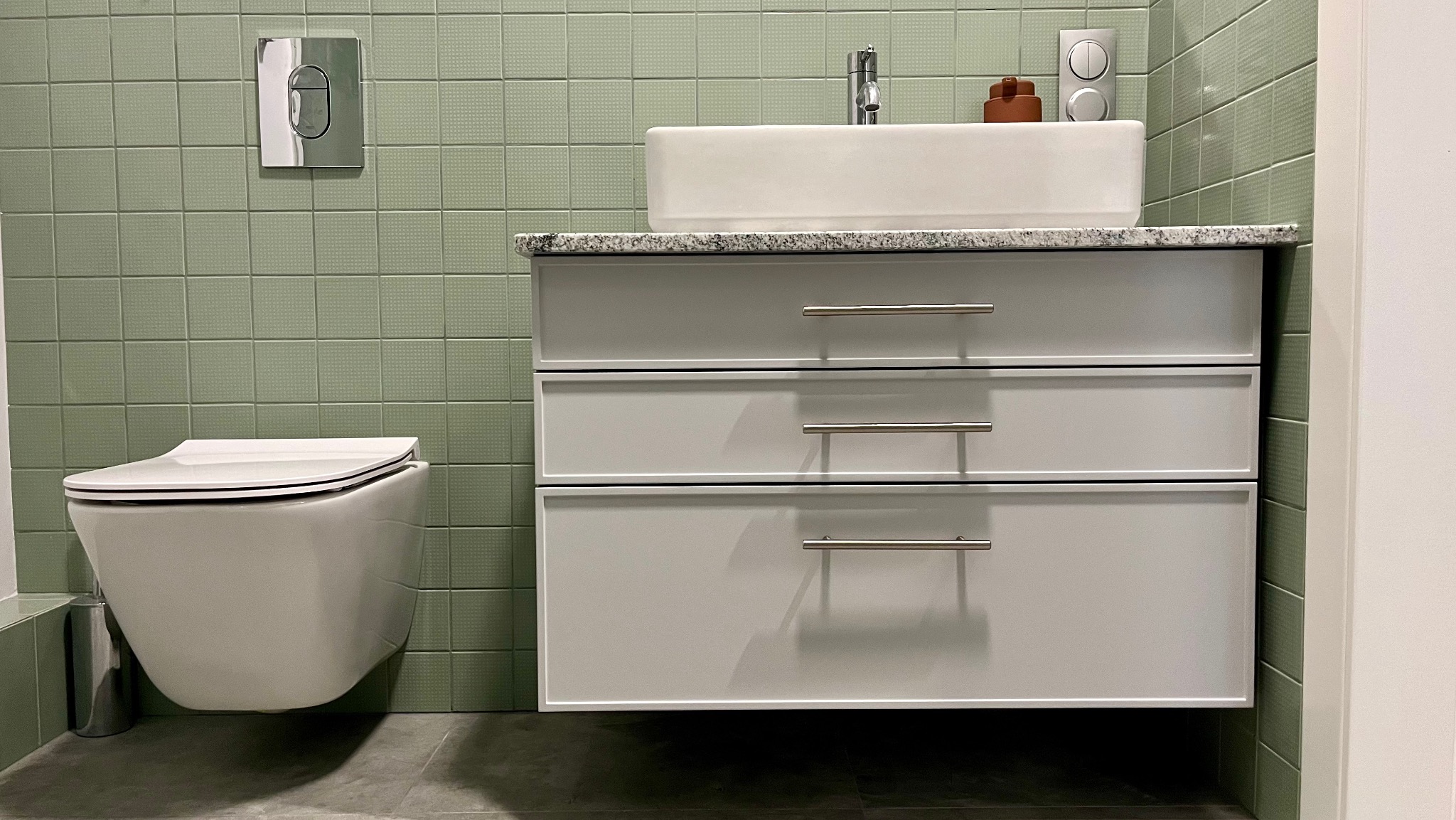 Nowoczesna łazienka z wiszącą toaletą i szafką pod umywalkę z trzema szufladami, wykończona zielonymi płytkami o delikatnej fakturze.