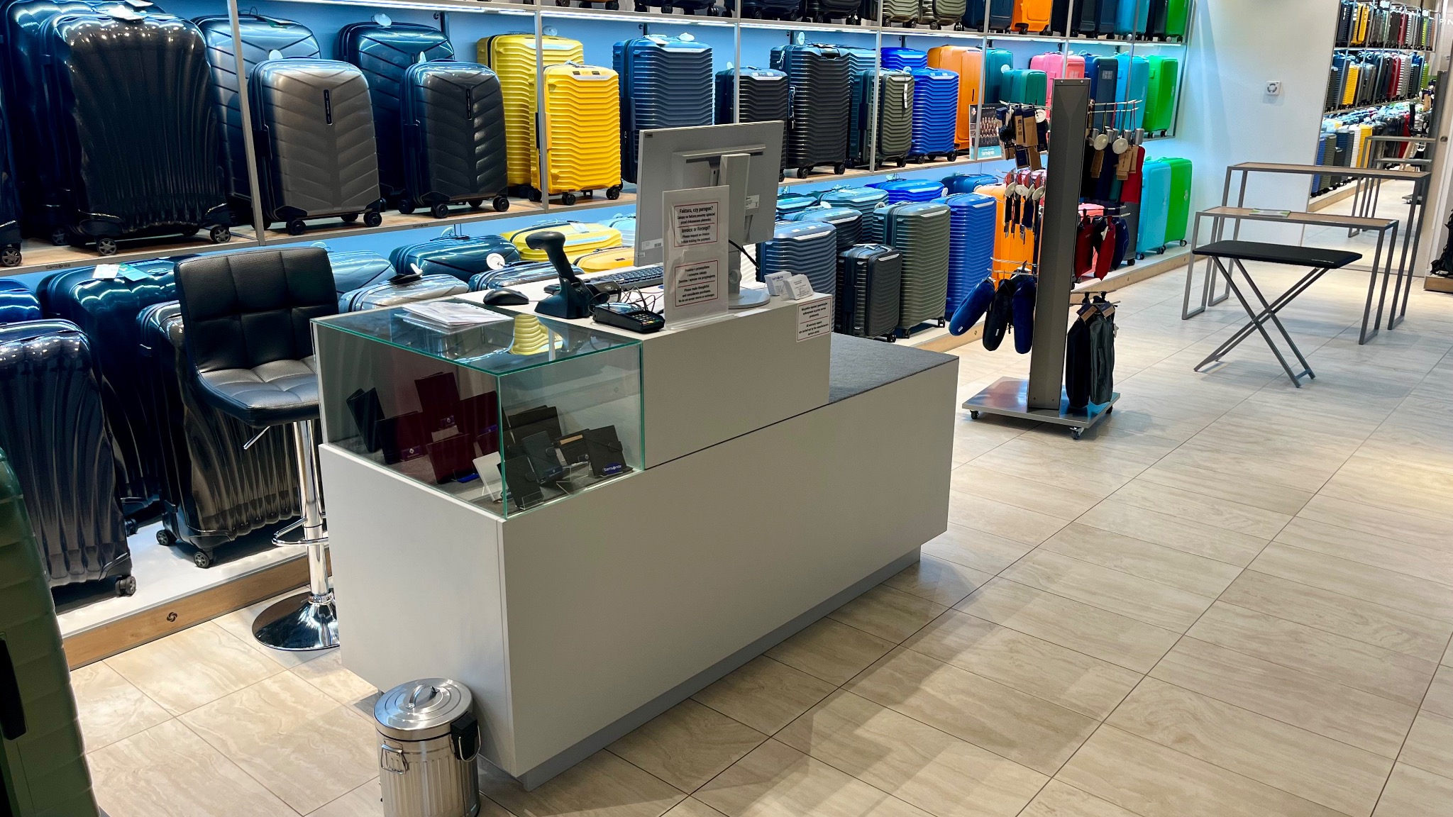 Wnętrze sklepu z szerokim wyborem kolorowych walizek na półkach, widoczna lada z komputerem i krzesłem barowym oraz ekspozycja akcesoriów podróżnych.