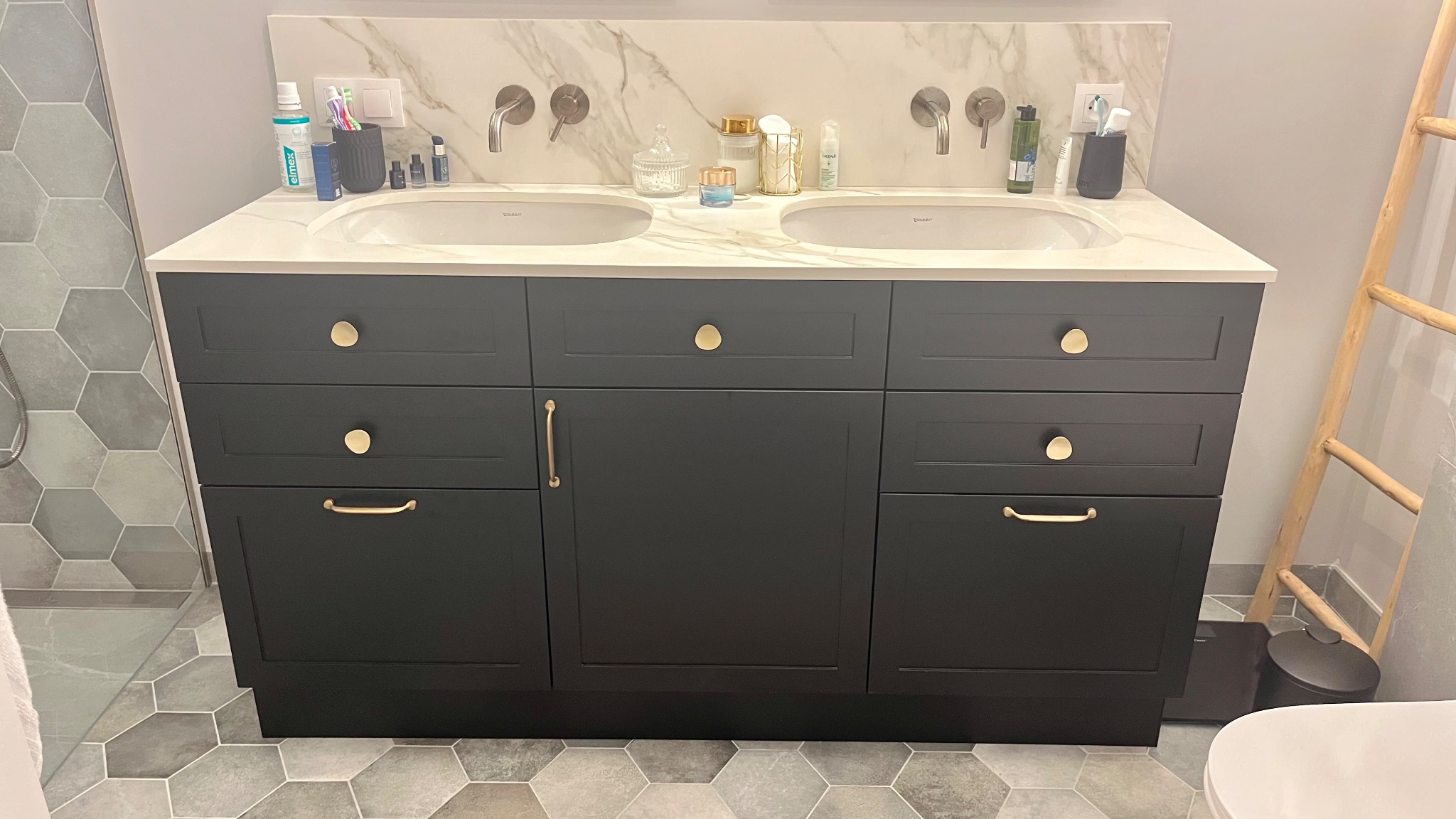 Czarna szafka łazienkowa z dwiema umywalkami, złotymi uchwytami i marmurowym blatem w nowoczesnej łazience z heksagonalnymi płytkami podłogowymi.