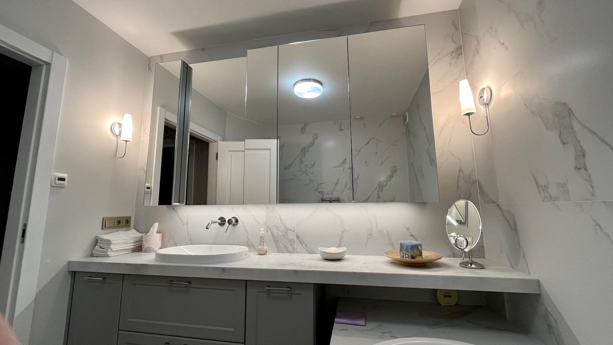 Nowoczesna łazienka z marmurową ścianą, umywalką nablatową, podświetlanym lustrem w szafce i minimalistycznymi kinkietami