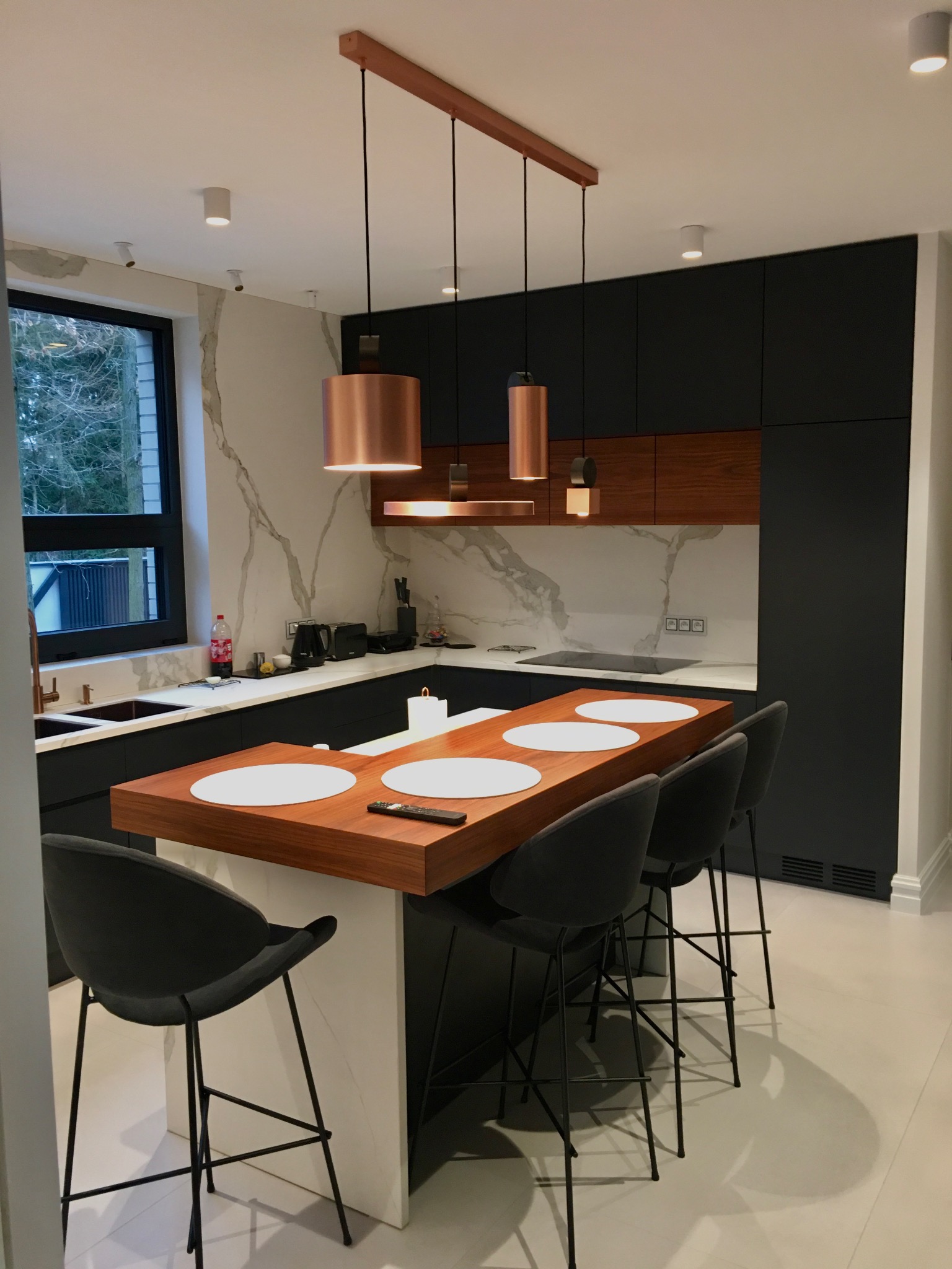 Nowoczesna kuchnia z wyspą, czarne szafki, drewniane akcenty, marmurowy backsplash i miedziane lampy wiszące nad blatem z hokerami.