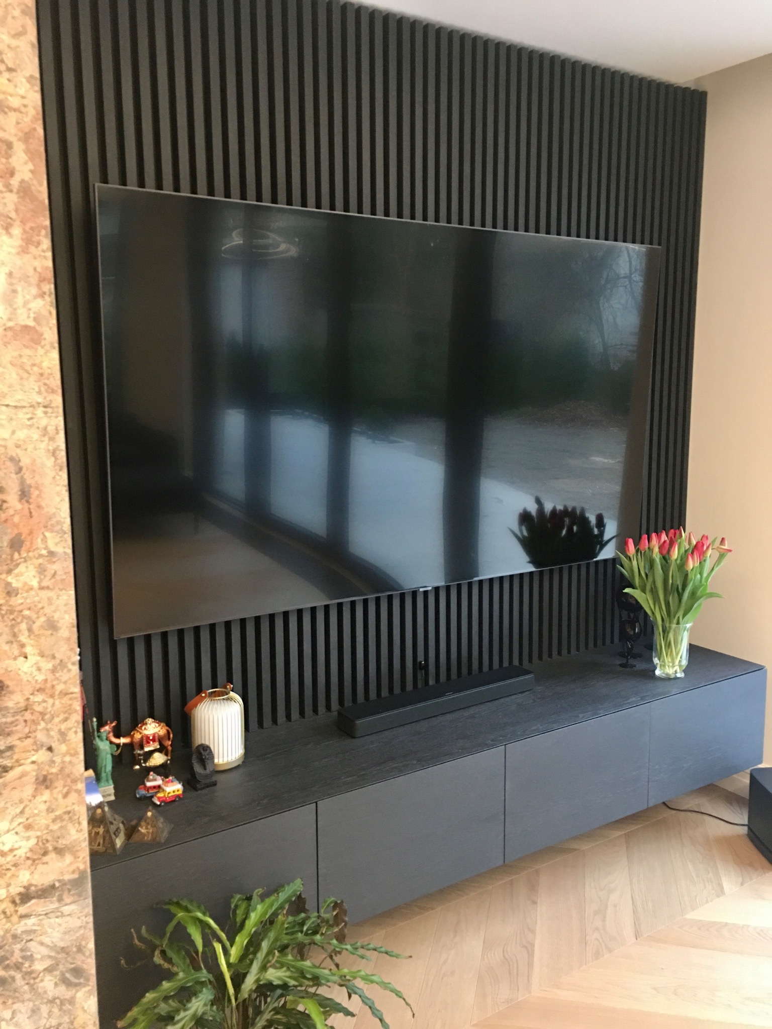 Ciemna szafka RTV z minimalistycznym frontem, zawieszona pod telewizorem na tle ściany z pionowych paneli dekoracyjnych, uzupełniona o bukiet tulipanów i drobne dekoracje.