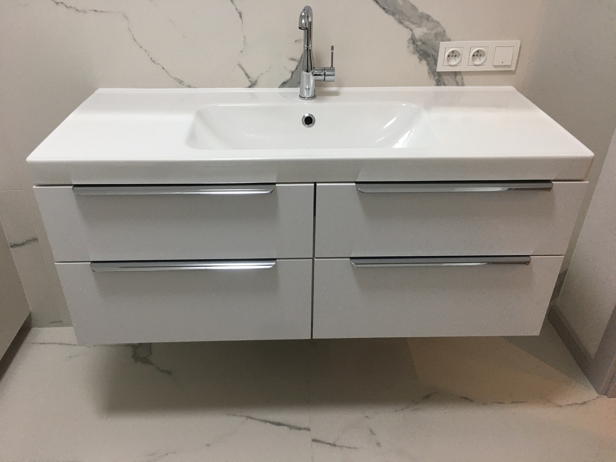 Biała umywalka z czterema szufladami i chromowanymi uchwytami, zamontowana na ścianie wyłożonej marmurowymi płytkami z szarymi żyłami, bateria umywalkowa w kolorze chromu.