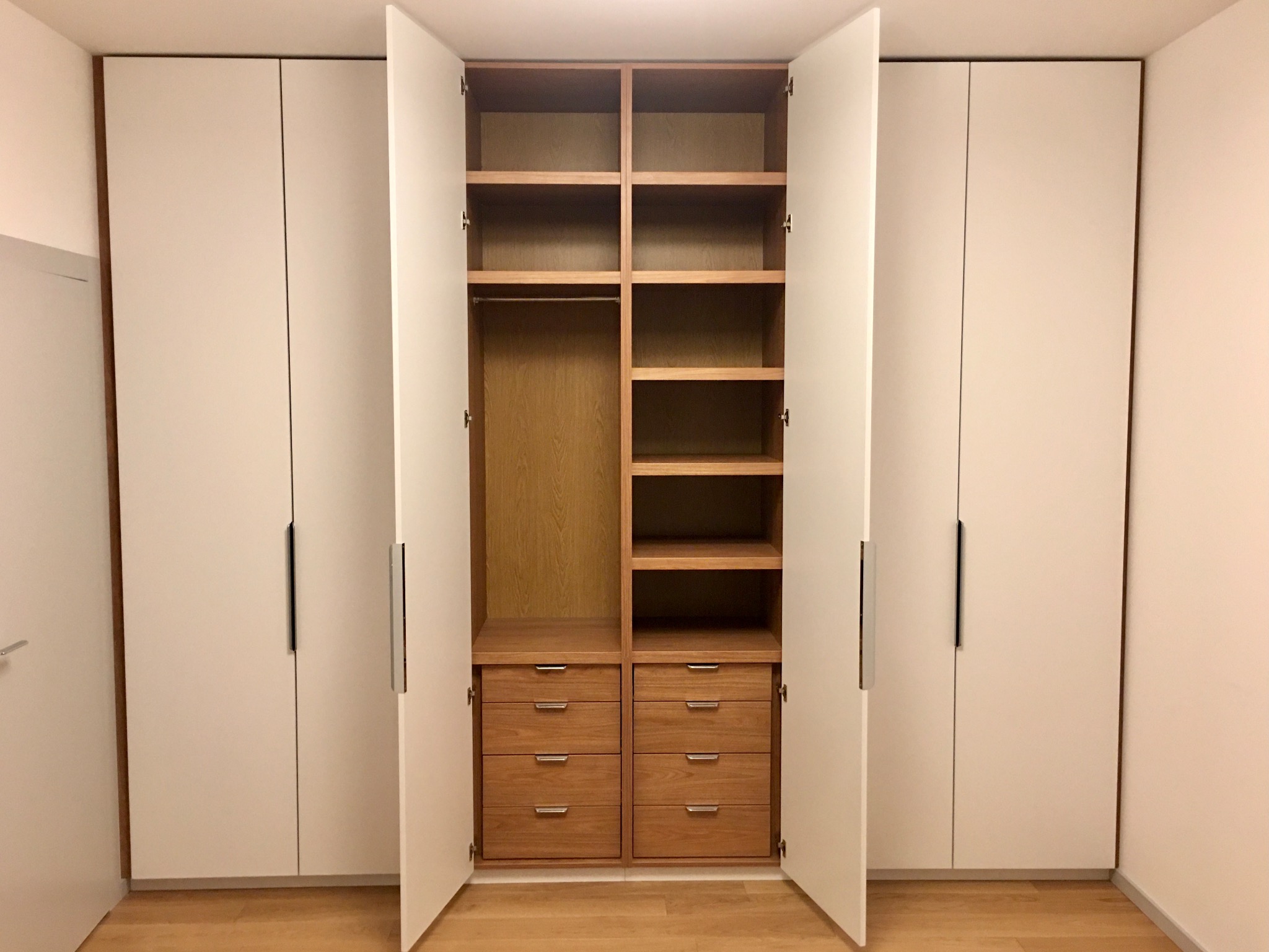 Biała szafa wnękowa z drewnianym wnętrzem i szufladami, otwarte drzwi ukazują półki i drążek na ubrania.