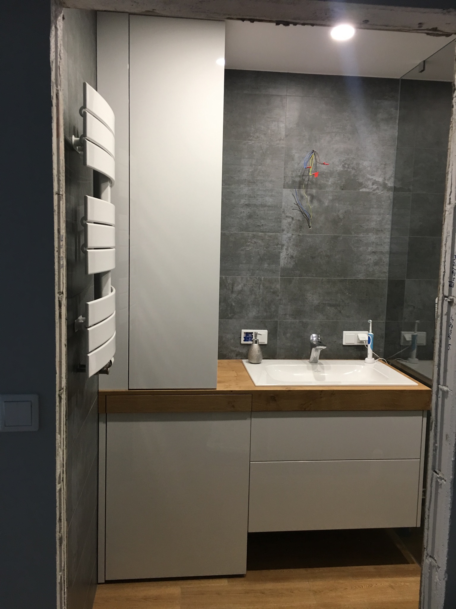 Nowoczesna łazienka w trakcie wykańczania: szare płytki, białe meble łazienkowe z drewnianym blatem, grzejnik drabinkowy i widoczne przewody elektryczne.