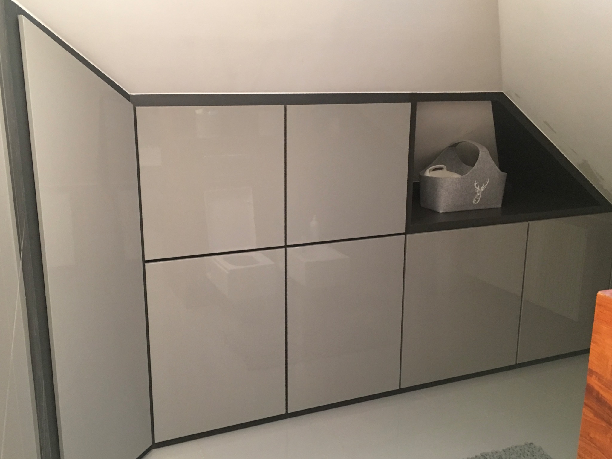 Jasnoszara zabudowa meblowa pod skosem, z otwartą półką i dekoracyjnym koszem z motywem jelenia.