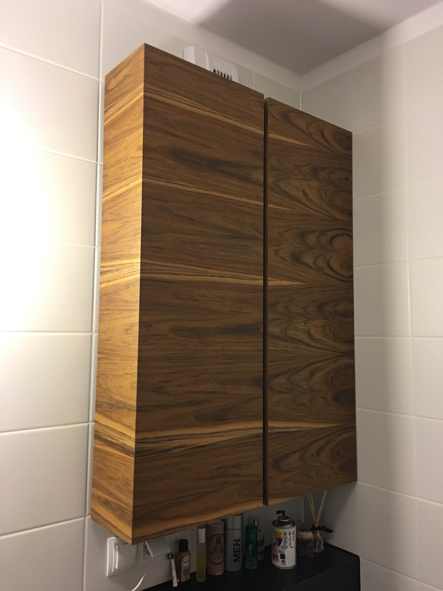 Wisząca szafka łazienkowa z forniru o wyraźnym usłojeniu, zamontowana na białych płytkach, pod spodem kosmetyki i dyfuzor zapachowy.