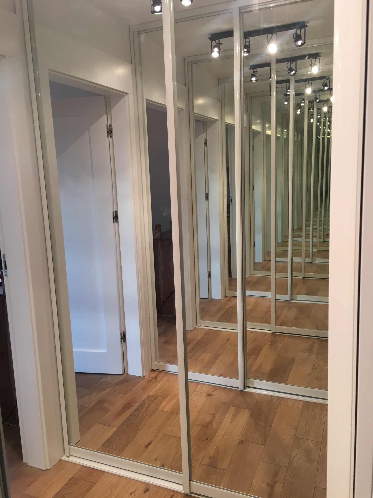 Długi korytarz z lustrzanymi drzwiami przesuwnymi, odbijającymi się w nieskończoność, ukazujący drewnianą podłogę i białe ościeżnice drzwiowe.