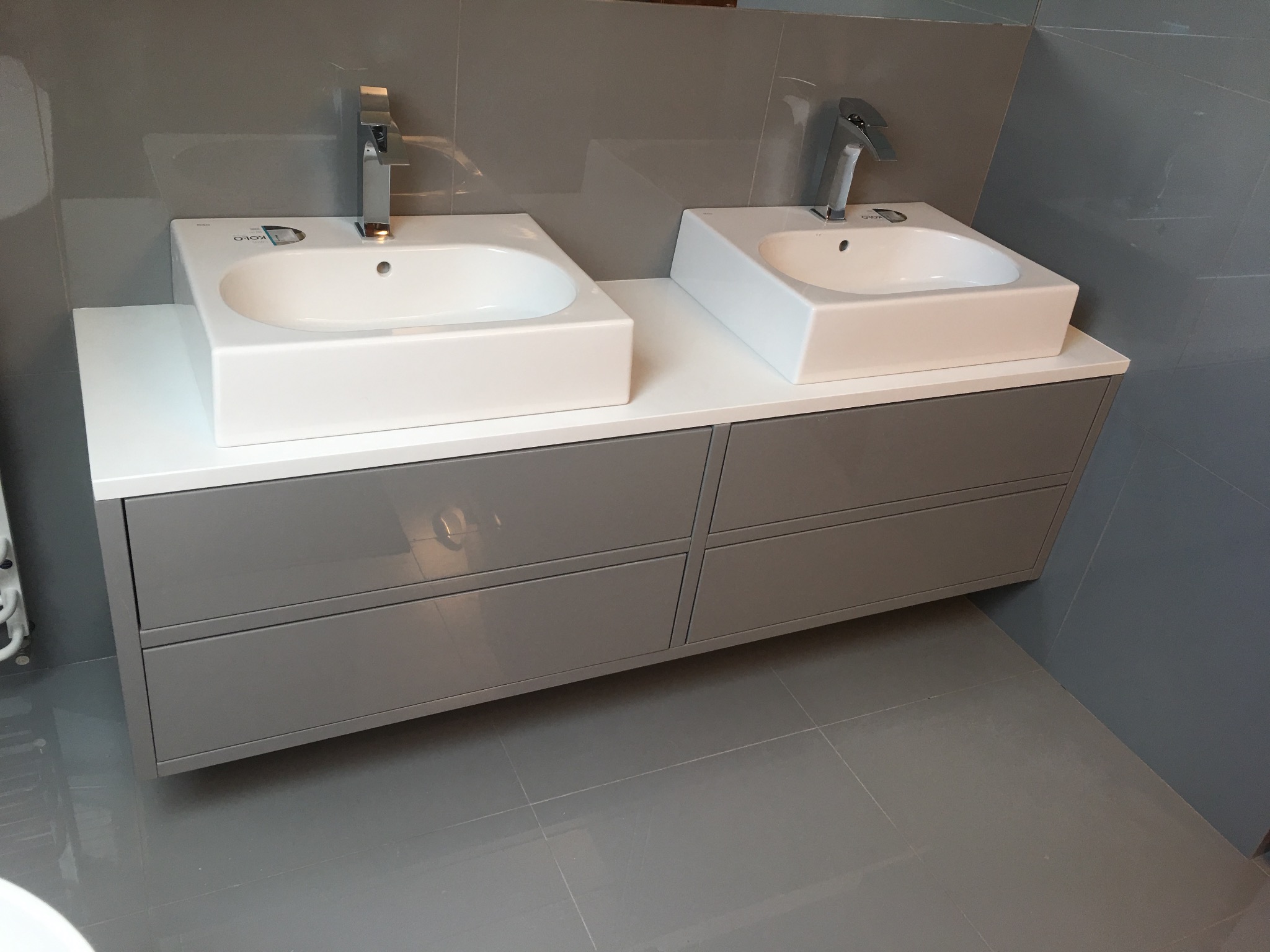 Dwie białe, prostokątne umywalki nablatowe z chromowanymi bateriami, osadzone na szarej szafce z czterema szufladami w nowoczesnej łazience z szarymi płytkami.