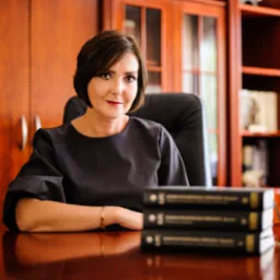 Portret kobiety w ciemnej sukience siedzącej za biurkiem z trzema czarnymi książkami, w tle drewniana biblioteczka.