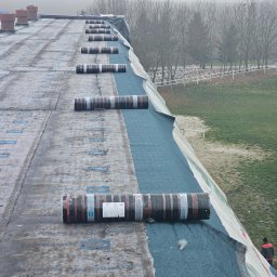 Zarzycki-System Paweł Zarzycki - Dach pokryty papą termozgrzewalną w rolkach, widoczne łączenia i częściowo ułożona warstwa, w tle zalesiony krajobraz w pochmurny dzień, na dachu widoczne kominy.