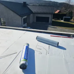 Biała membrana dachowa RESITRIX SKW w rolce na dachu pokrytym płytami izolacyjnymi, w tle dom z ciemną blachą na dachu i zalesione wzgórza.