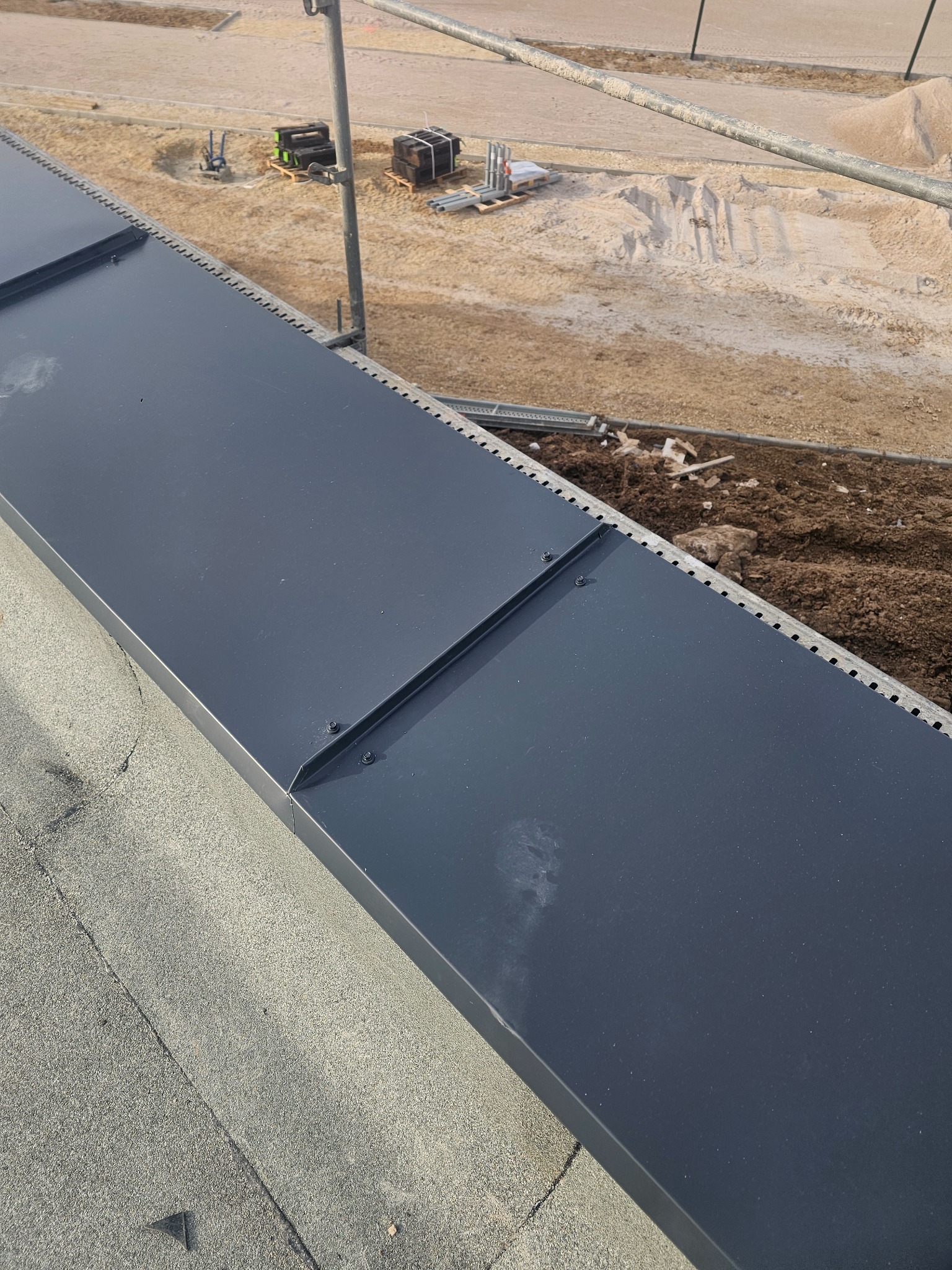 Nowe, ciemnoszare panele dachowe z widocznymi śrubami mocującymi, ułożone na papie bitumicznej, z placem budowy w tle.