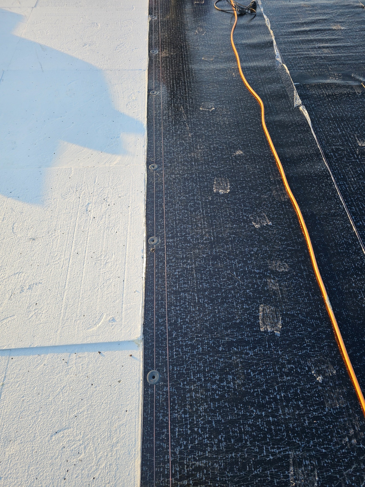 Biała izolacja dachu styropianem, połączona z czarną membraną dachową przy pomocy metalowych podkładek i sznurków, pomarańczowy kabel zasilający leżący na membranie.