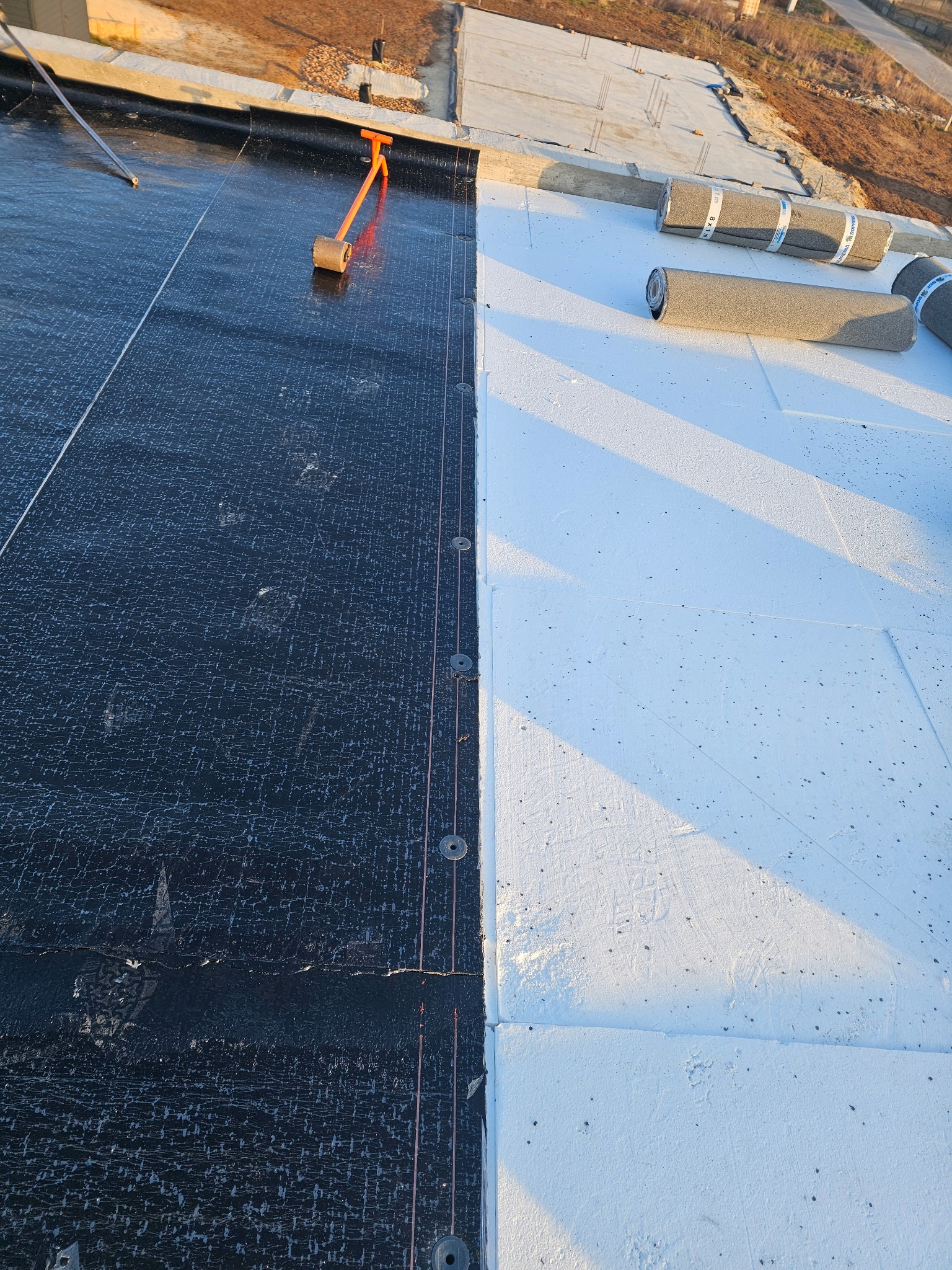 Fragment dachu płaskiego w trakcie prac: po lewej czarna papa z widocznymi śladami stóp i narzędziem do jej mocowania, po prawej białe płyty izolacyjne, w tle rolki papy. Widoczne łączenie papy...