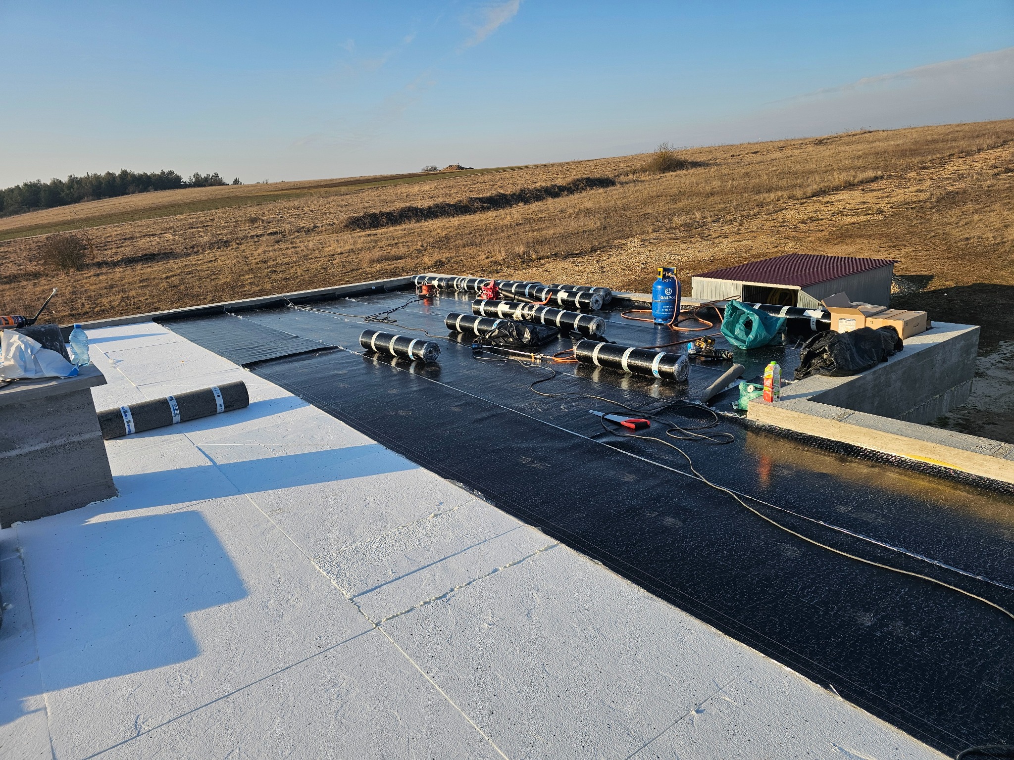 Prace na dachu: rolki papy termozgrzewalnej ułożone na czarnej membranie, butla gazowa, narzędzia, widok na pagórkowaty teren w słoneczny dzień.