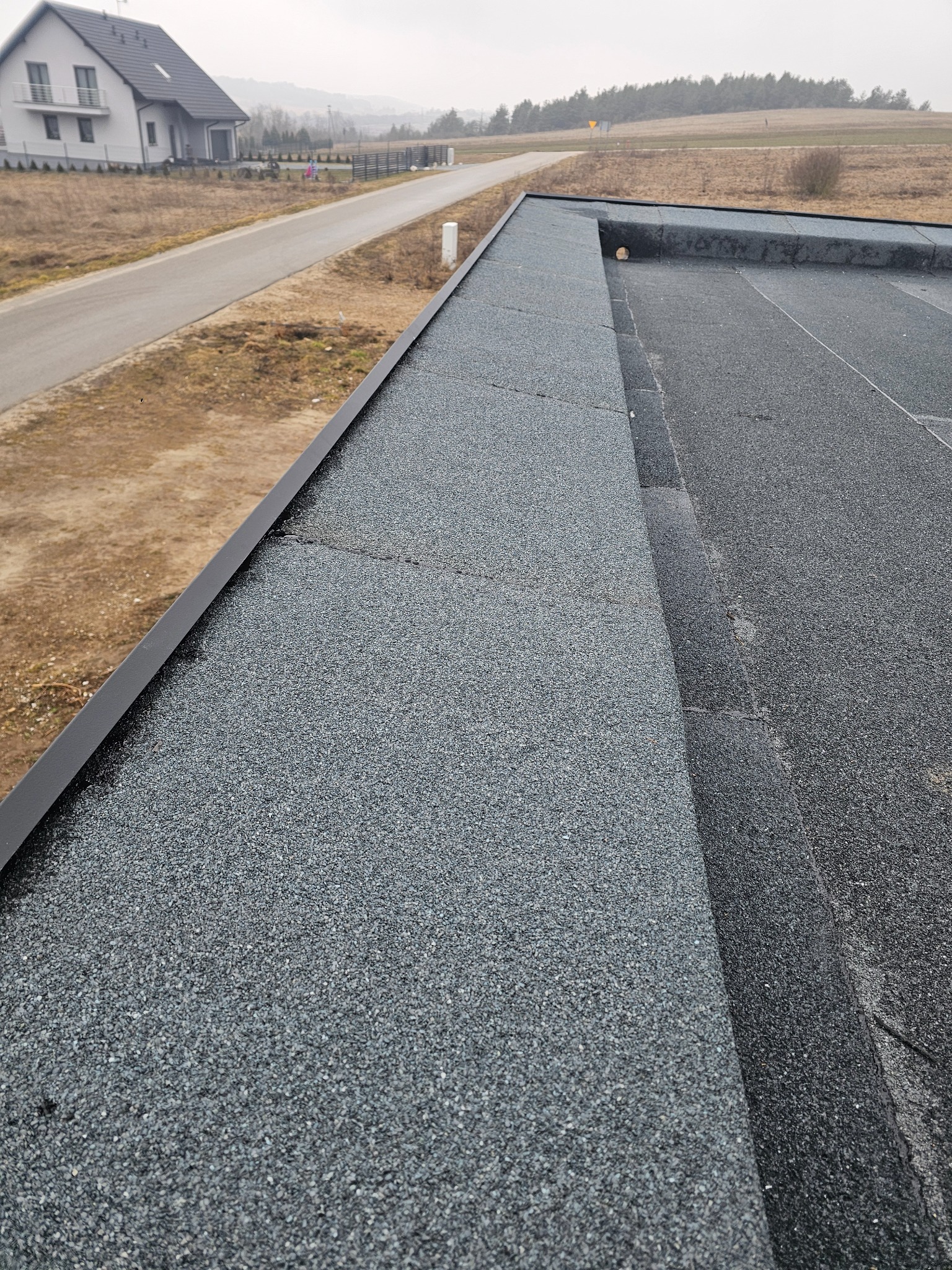 Widok z góry na płaski dach pokryty papą z widocznym systemem odprowadzania wody i obróbką blacharską, w tle nowy dom jednorodzinny i krajobraz.