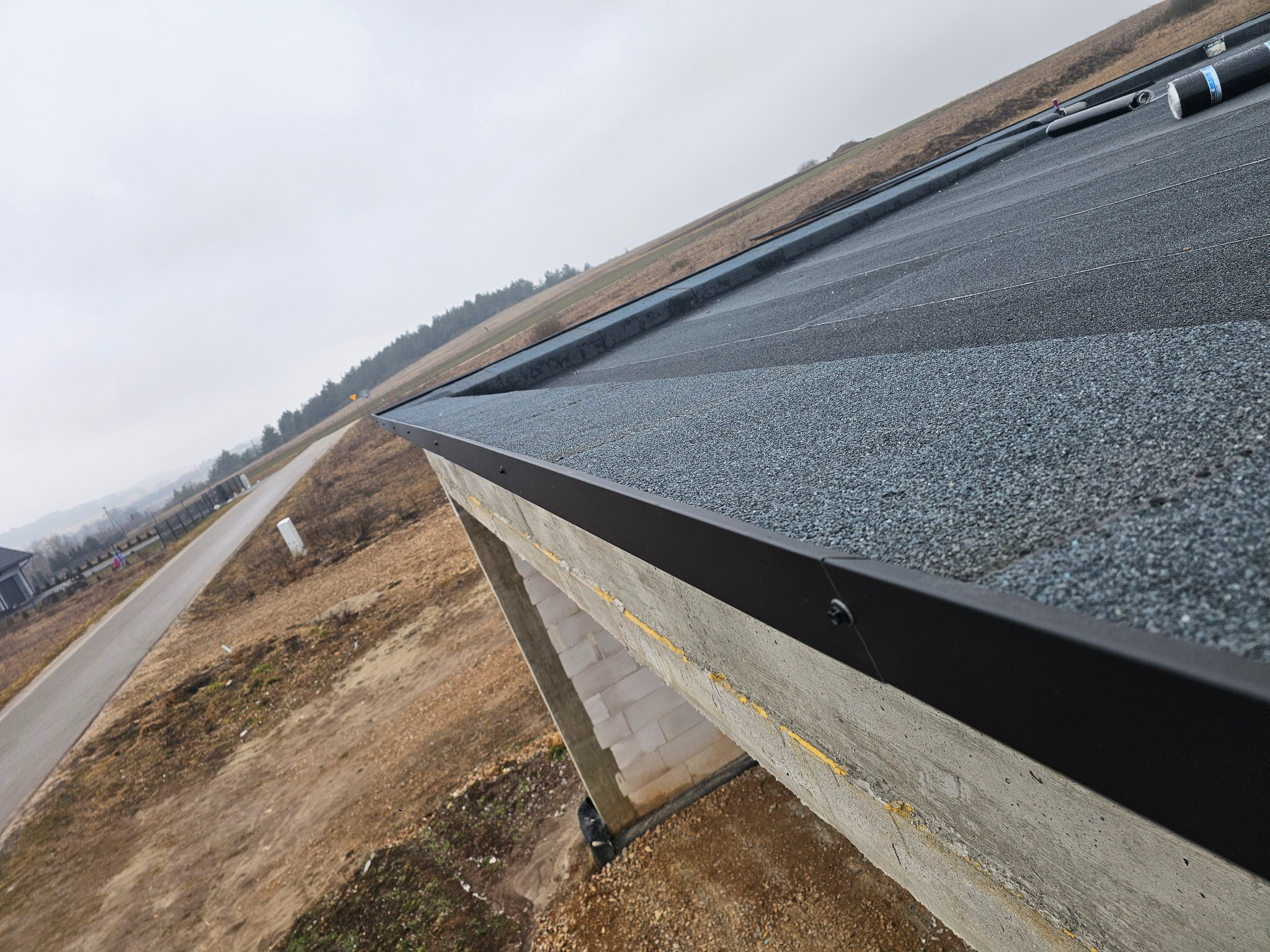 Widok z góry na płaski dach pokryty papą z posypką, z widoczną obróbką blacharską krawędzi, w tle droga i pola w pochmurny dzień.