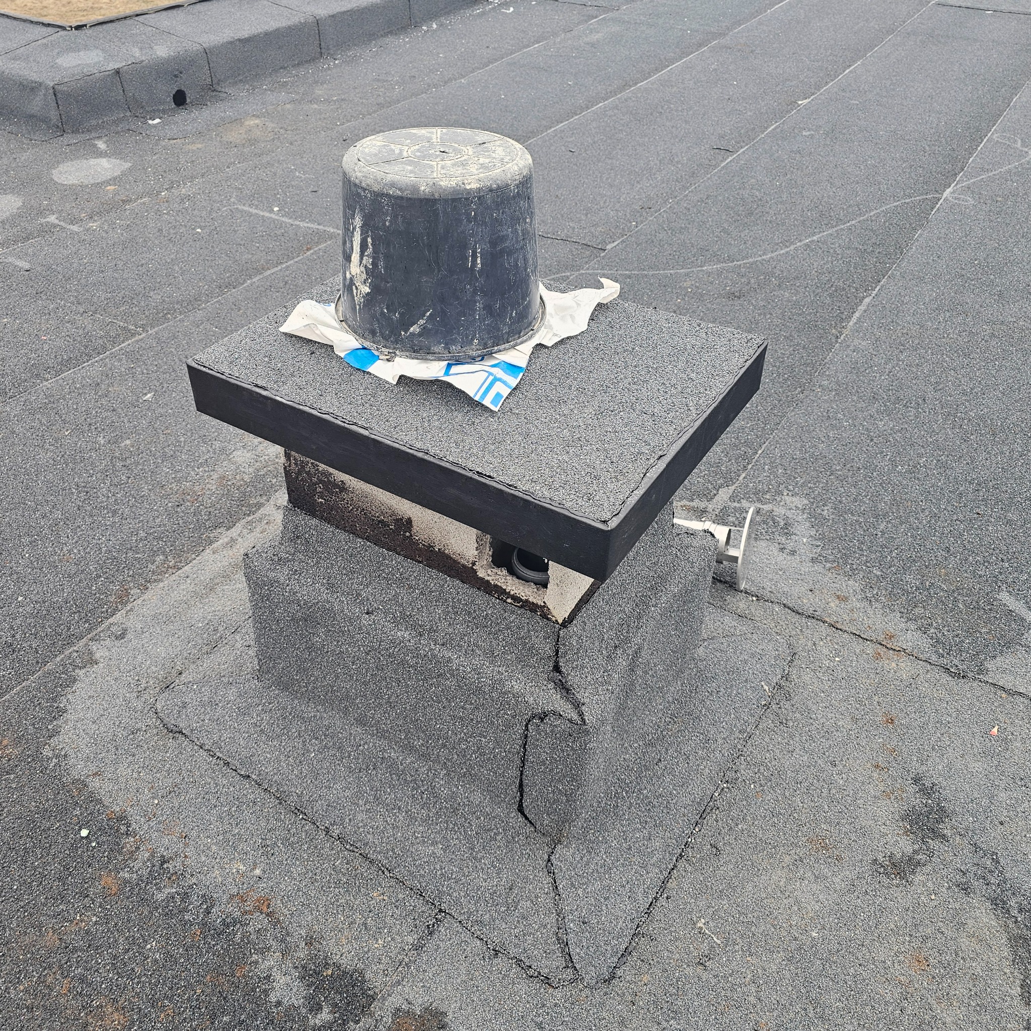 Płaski dach pokryty papą z zabudowanym kominkiem wentylacyjnym i czarnym, brudnym wiadrem na jego szczycie, fragmenty uszkodzeń na papie