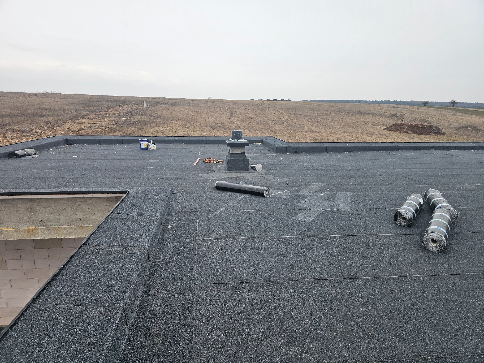 Widok z góry na płaski dach pokryty czarną papą, z widocznymi rolkami papy, kominem wentylacyjnym i narzędziami, w tle rozległe pole pod pochmurnym niebem.