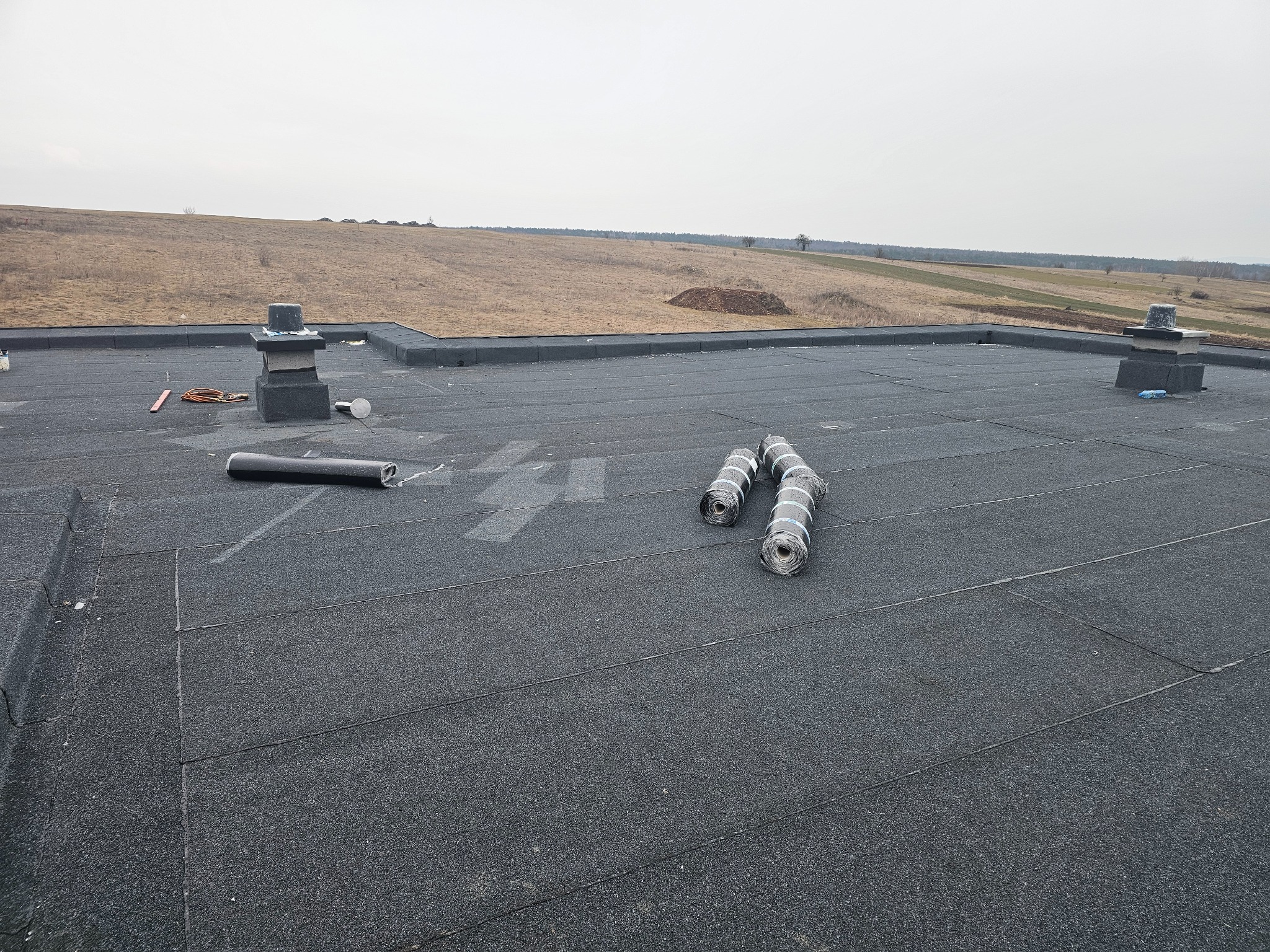 Widok z góry na płaski dach pokryty papą, z widocznymi kominami i rolkami materiału izolacyjnego, tło stanowi pagórkowaty krajobraz pod pochmurnym niebem.