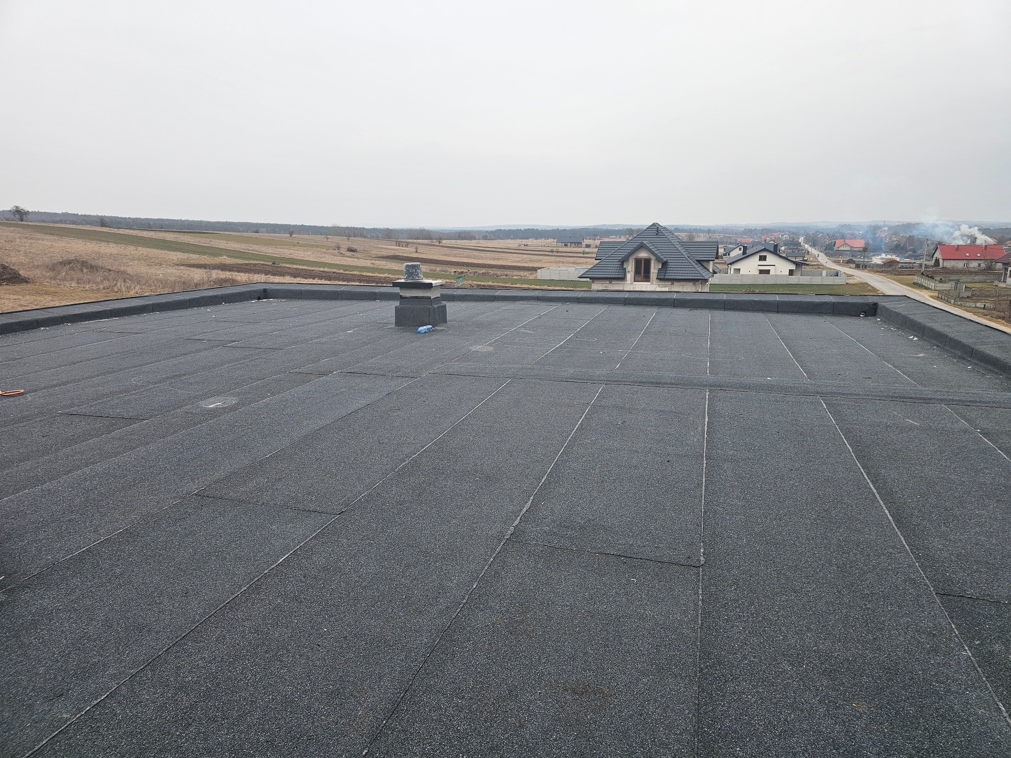 Czarny, płaski dach pokryty papą termozgrzewalną z widocznym kominem wentylacyjnym i krajobrazem wiejskim w tle.