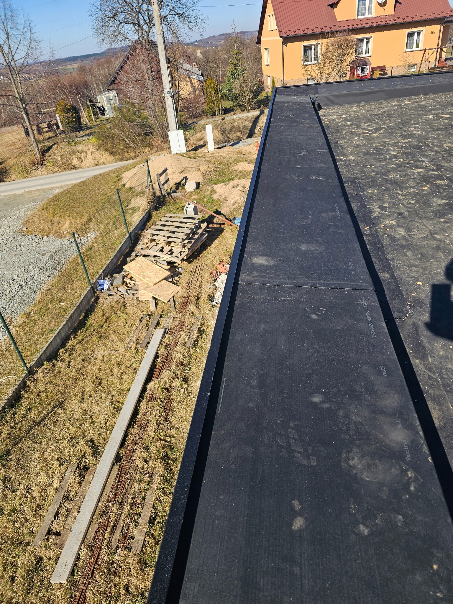 Widok z góry na płaski dach pokryty czarną papą termozgrzewalną, z widocznymi łączeniami; w tle budynek mieszkalny i teren z materiałami budowlanymi.