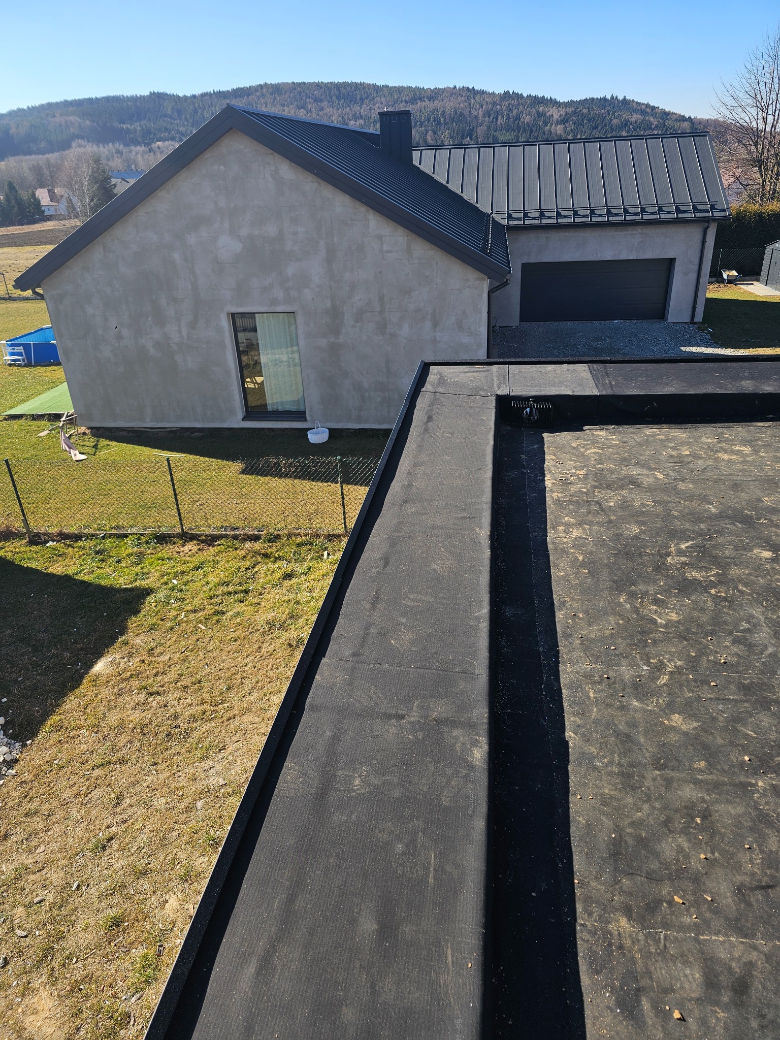 Czarna, płaska powierzchnia dachu pokryta papą termozgrzewalną, widoczny odpływ wody deszczowej, w tle dom jednorodzinny z szarą blachodachówką i zielony trawnik z basenem.