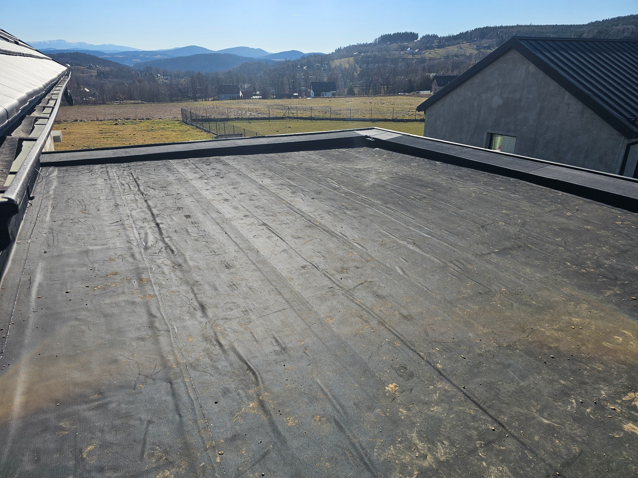 Wykończony płaski dach pokryty czarną papą termozgrzewalną z widocznymi zgrzewami, na tle krajobrazu z pagórkami i zabudowaniami.
