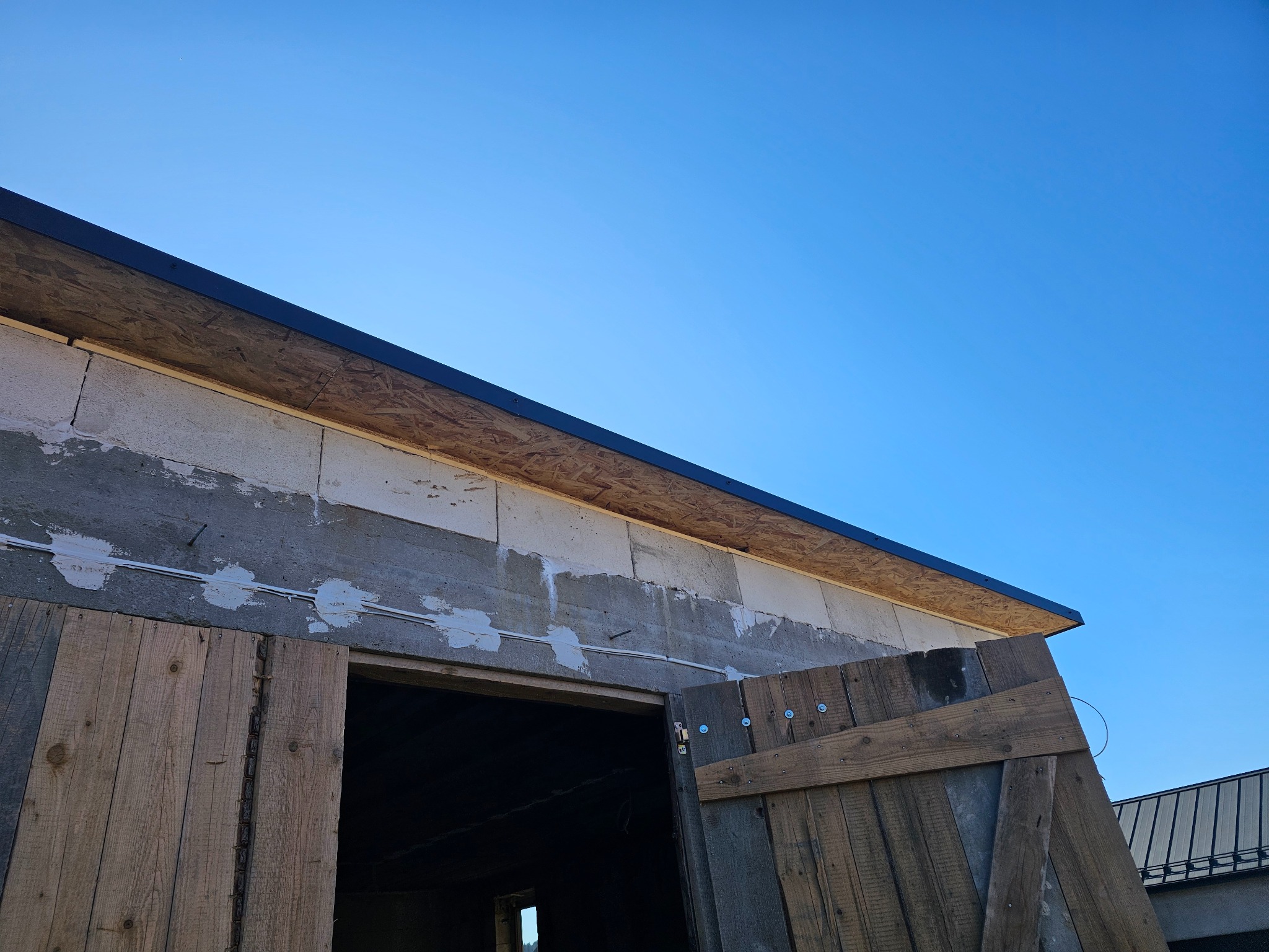 Fragment budynku z drewnianymi drzwiami w trakcie budowy, widoczne bloczki betonowe, płyta OSB pod dachem i czarny pas blachy na krawędzi dachu na tle błękitnego nieba.