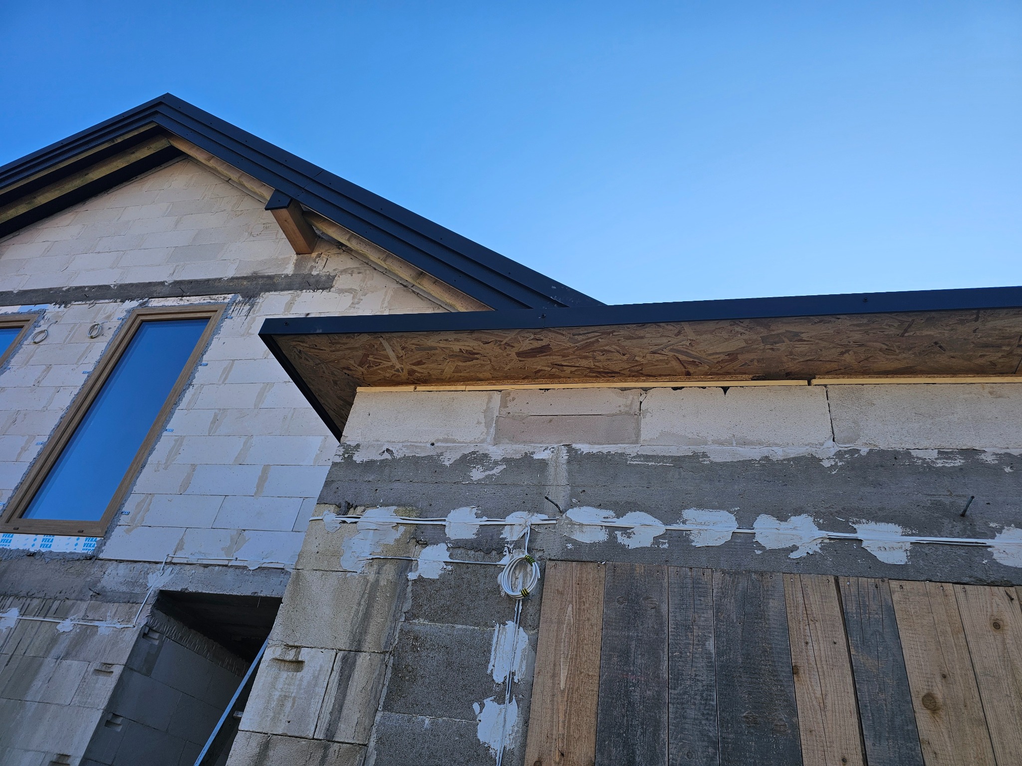 Fragment niedokończonego domu z widocznym dachem z czarną blachodachówką, oknem dachowym i ścianami z betonu komórkowego w trakcie budowy.