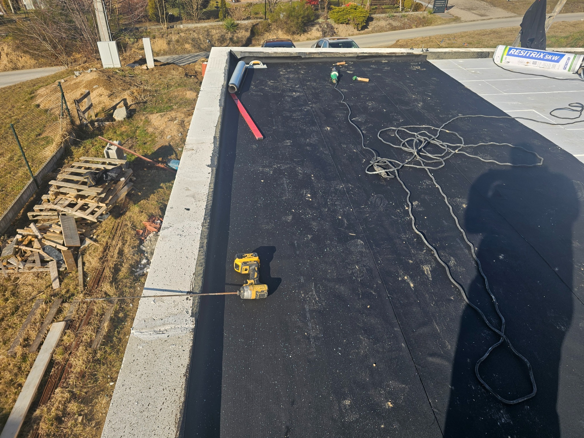 Widok z góry na płaski dach pokryty czarną membraną bitumiczną, w trakcie prac dekarskich. Widoczne narzędzia: wkrętarka akumulatorowa, zwinięta rolka papy, poziomica i opakowanie po materiale...