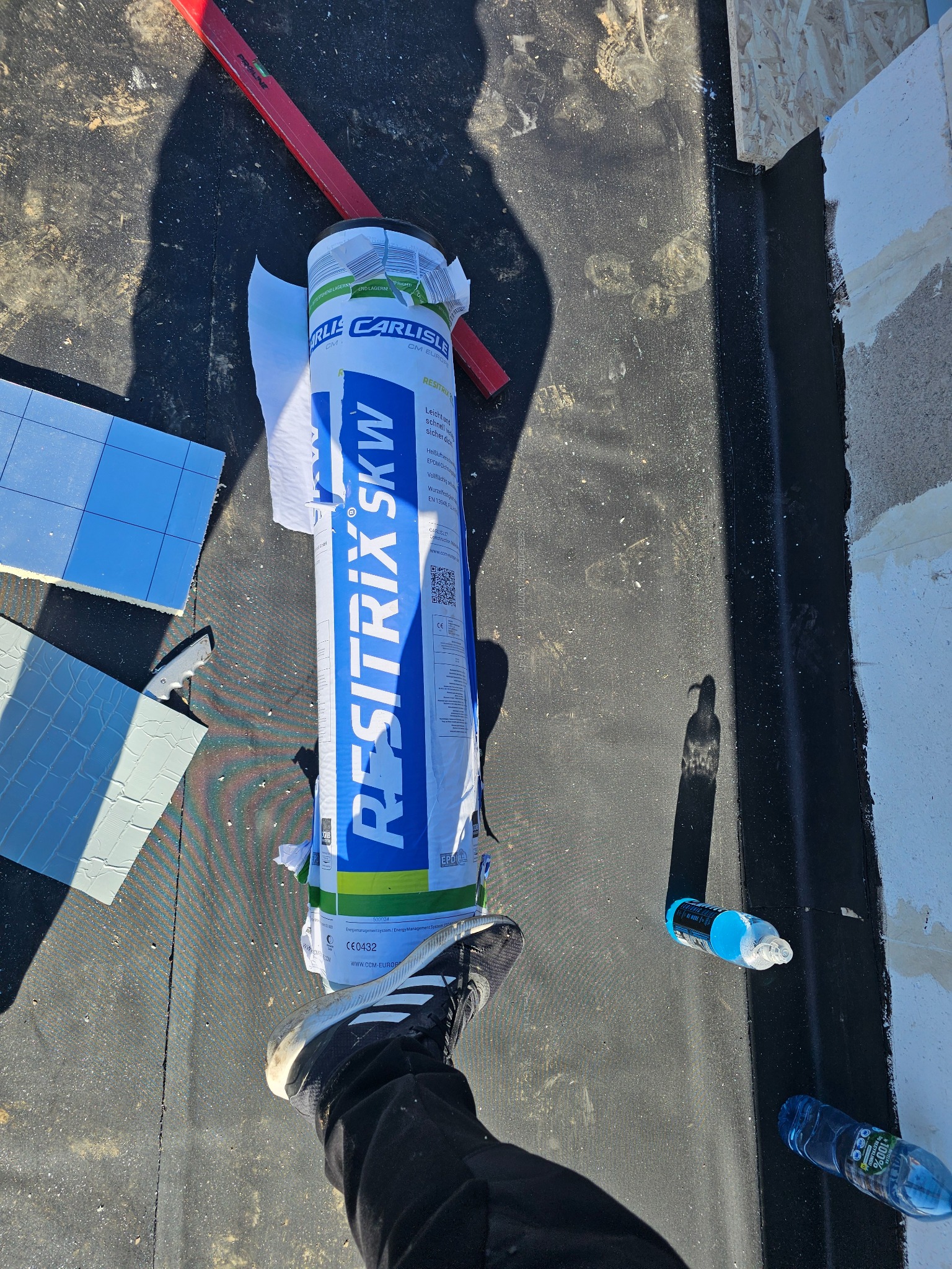 Rolka membrany dachowej Resitrix SK W na czarnym dachu z widocznym fragmentem nogi w czarnym obuwiu sportowym, poziomica, fragmenty termoizolacji, nóż dekarski i dwie butelki z wodą.