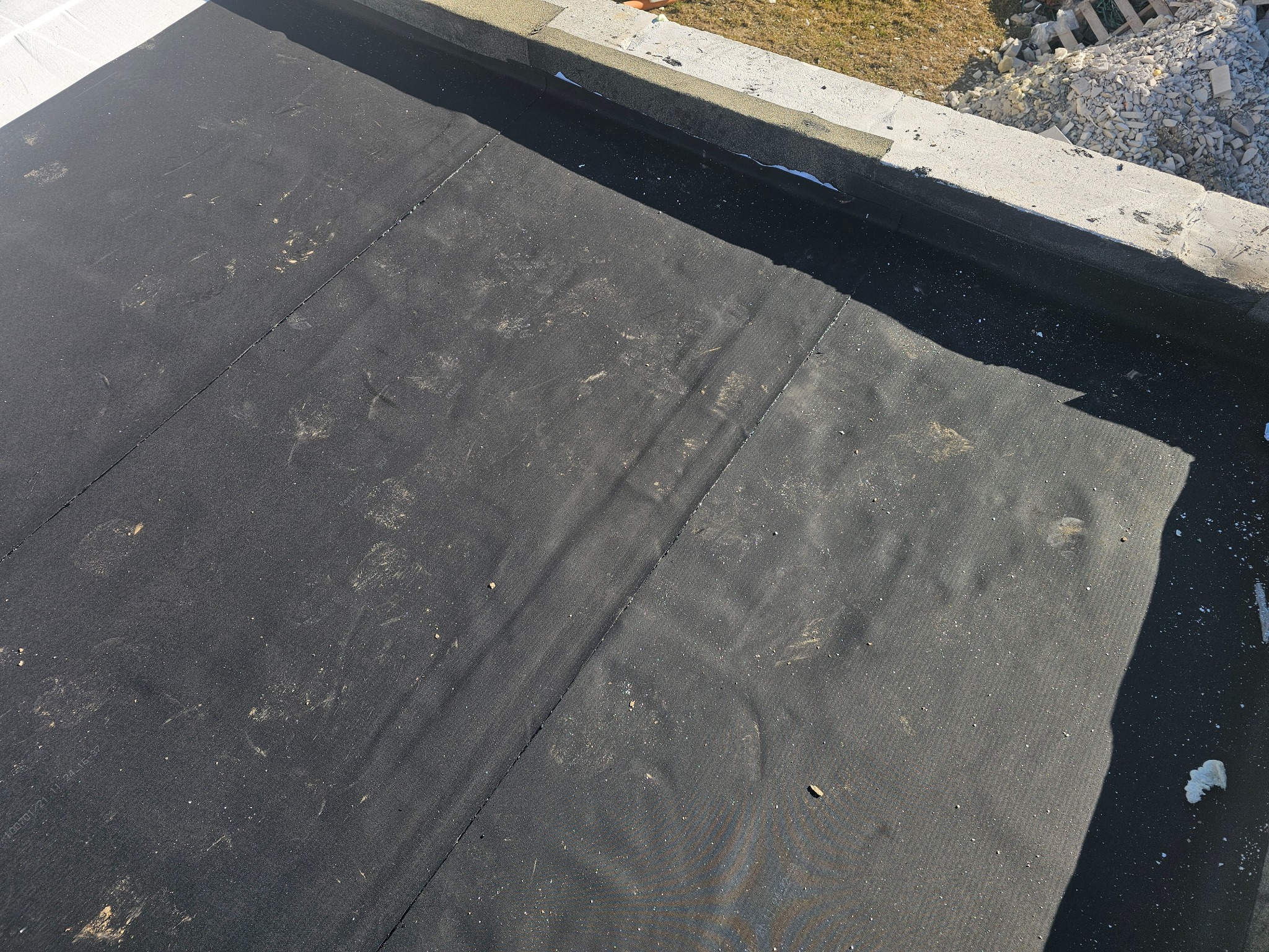 Fragment pokrycia dachu papą termozgrzewalną z widocznymi śladami stóp i datą produkcji na powierzchni, obrzeże dachu wykończone betonowymi elementami