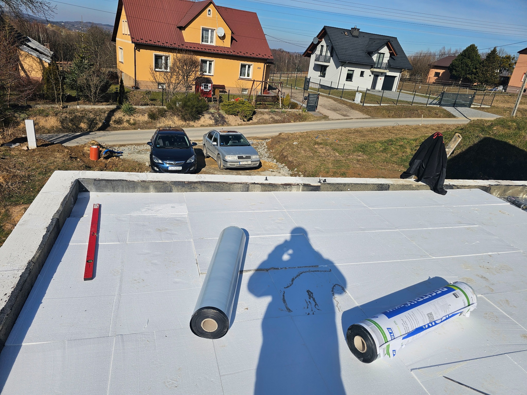 Na dachu budynku w trakcie prac izolacyjnych leżą rolki papy termozgrzewalnej, poziomica i widać cień osoby wykonującej zdjęcie; w tle dwa domy i samochody.