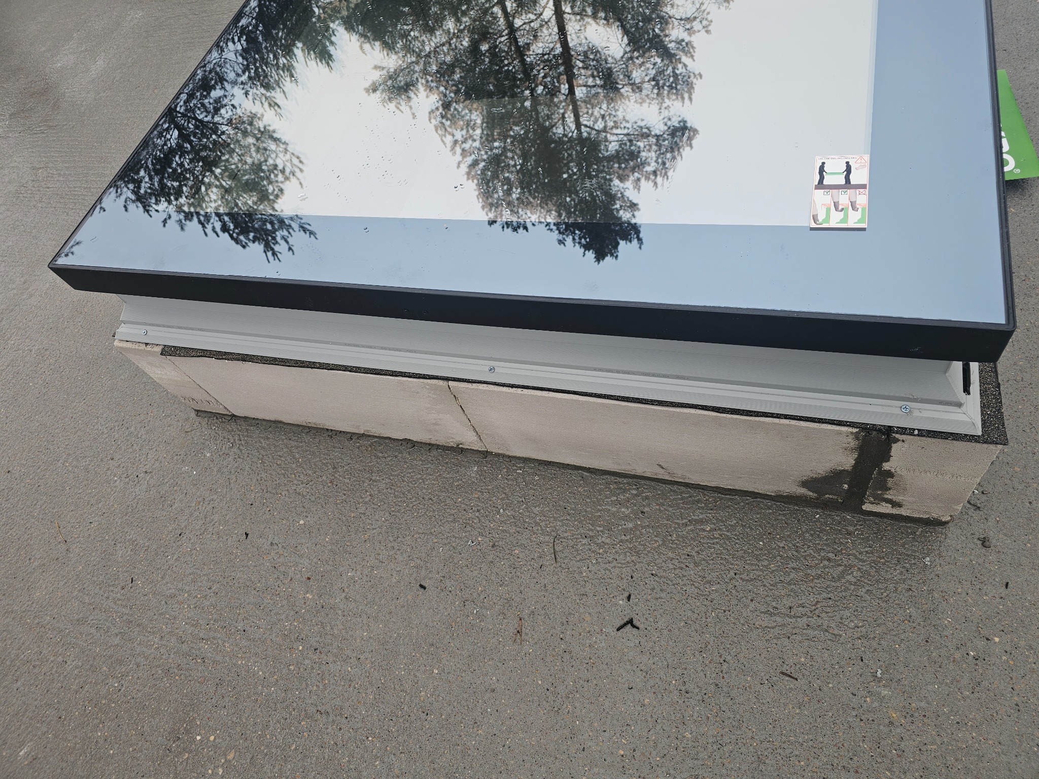 Nowo zamontowany świetlik dachowy z czarną ramą, widoczne odbicie drzew na szkle, uszczelnienie na styku z dachem.