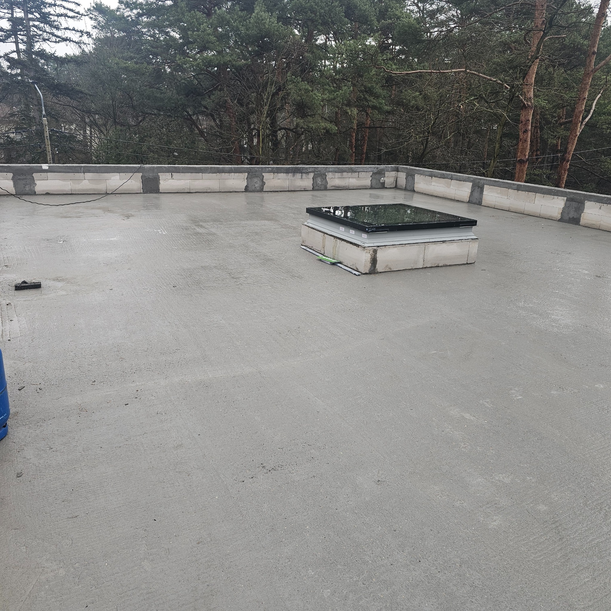 Wykończony, płaski dach pokryty szarą, gładką wylewką betonową z zamontowanym świetlikiem dachowym. Widoczne bloczki betonowe na obrzeżach dachu i otaczający las.